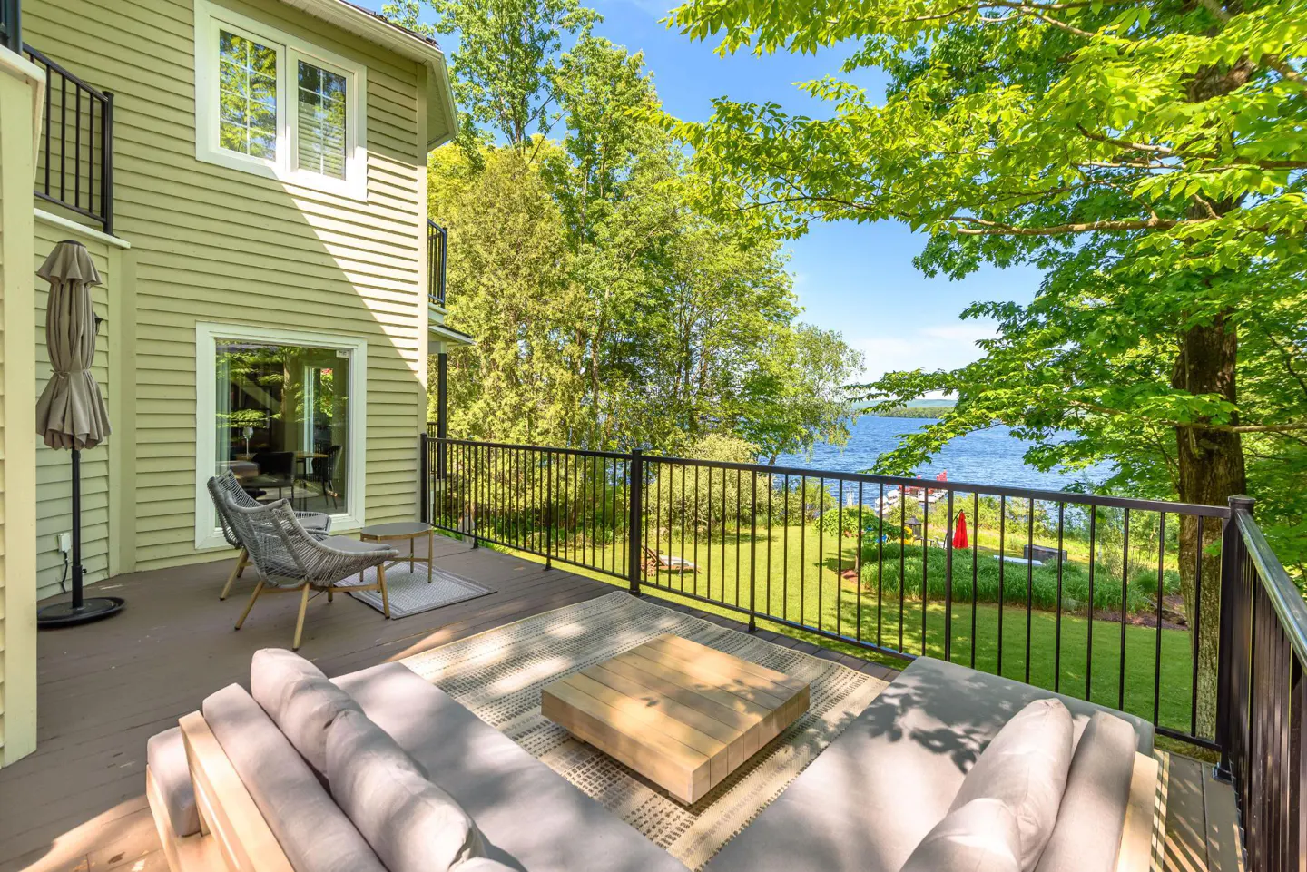 Outdoor deck with gray sofa, wood table, and chairs overlooking a green lawn and blue lake on a sunny day.