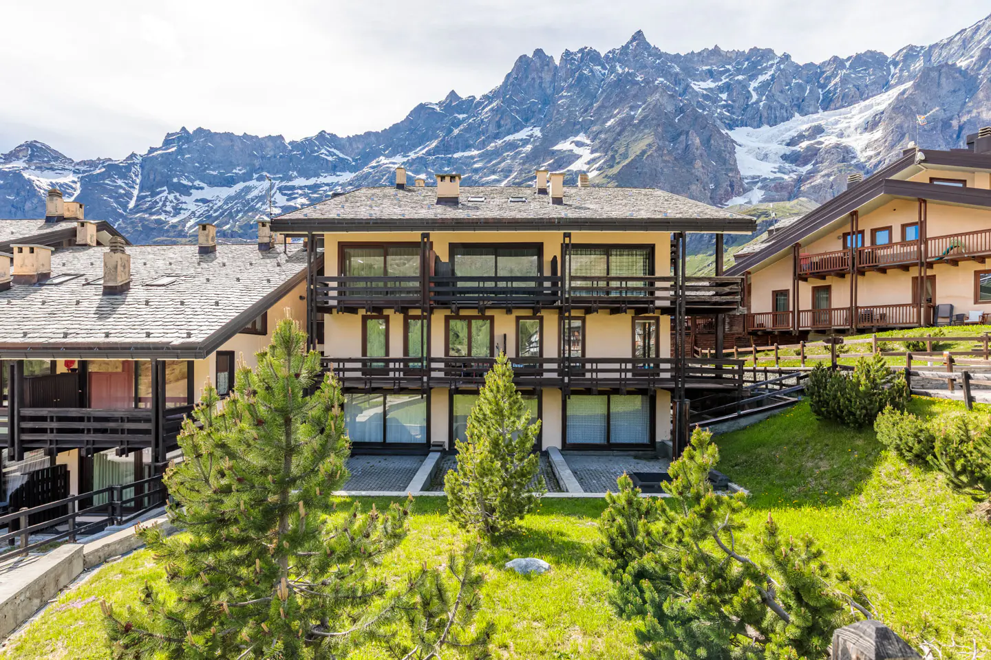 A multi-story chalet with dark wood balconies sits on a grassy slope before a backdrop of snow-capped mountain peaks.