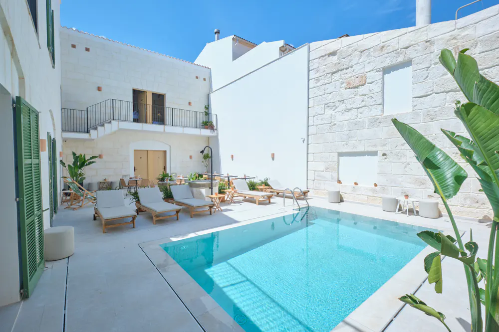 Outdoor pool area with lounge chairs, white stone walls, and green shutters under a clear blue sky.