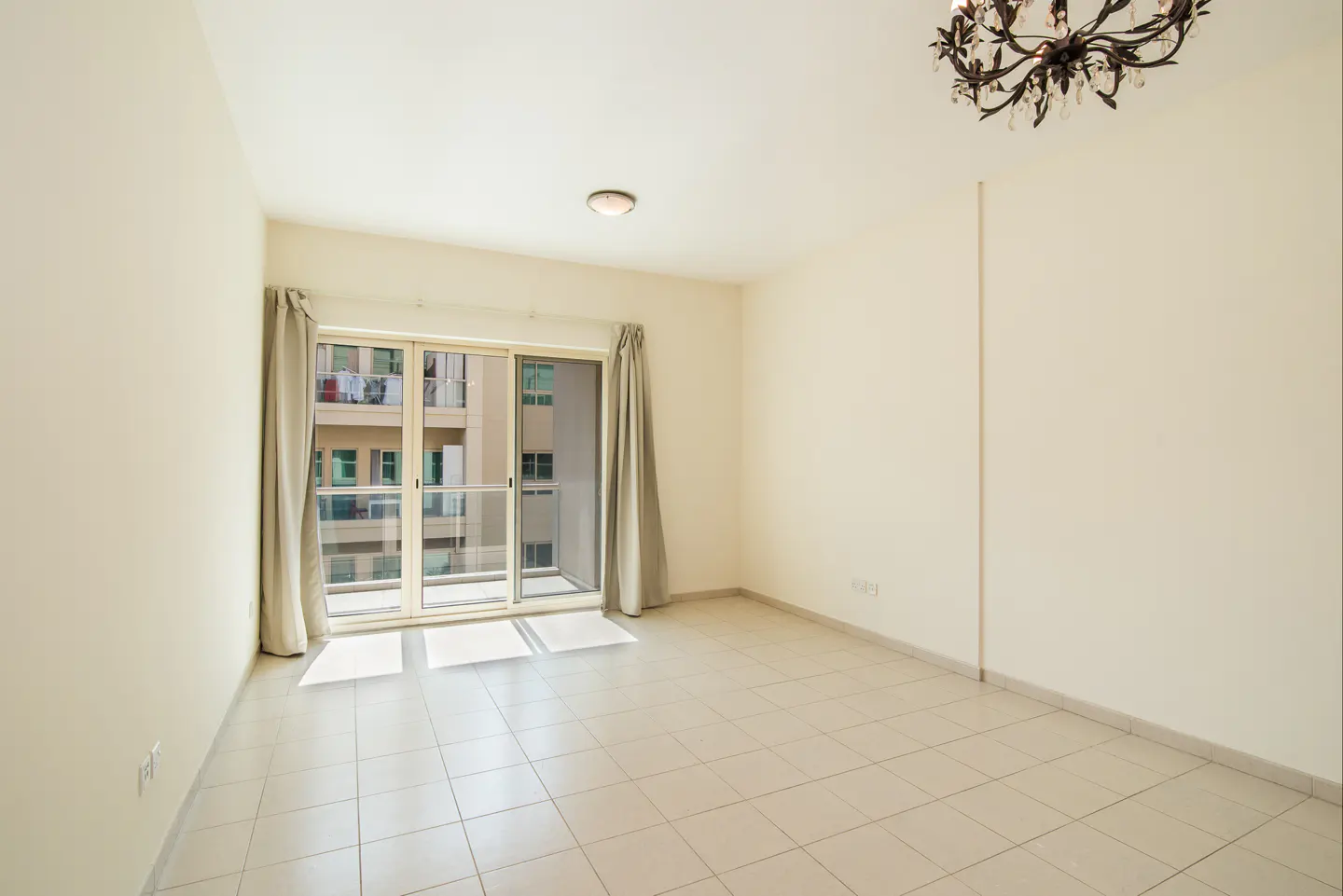 Empty room with beige walls, tiled floor, and a chandelier. Sliding glass doors lead to a balcony with a view of another building.