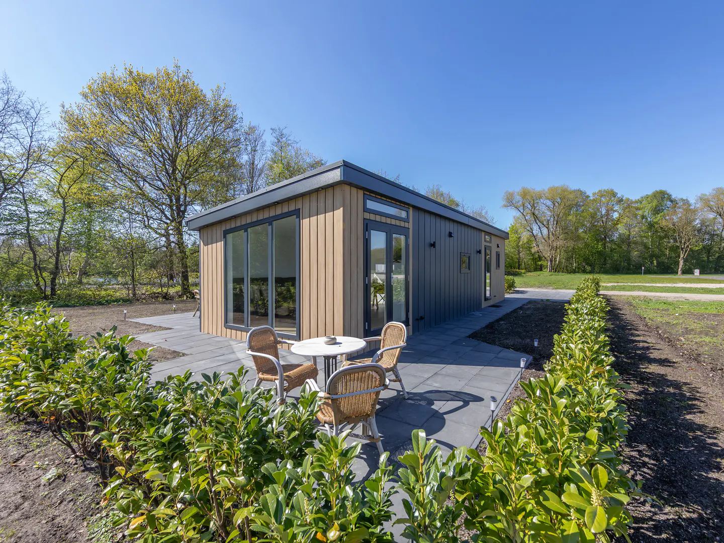 Modern home with wood and gray siding, a patio with a table and chairs, and green landscaping under a blue sky.