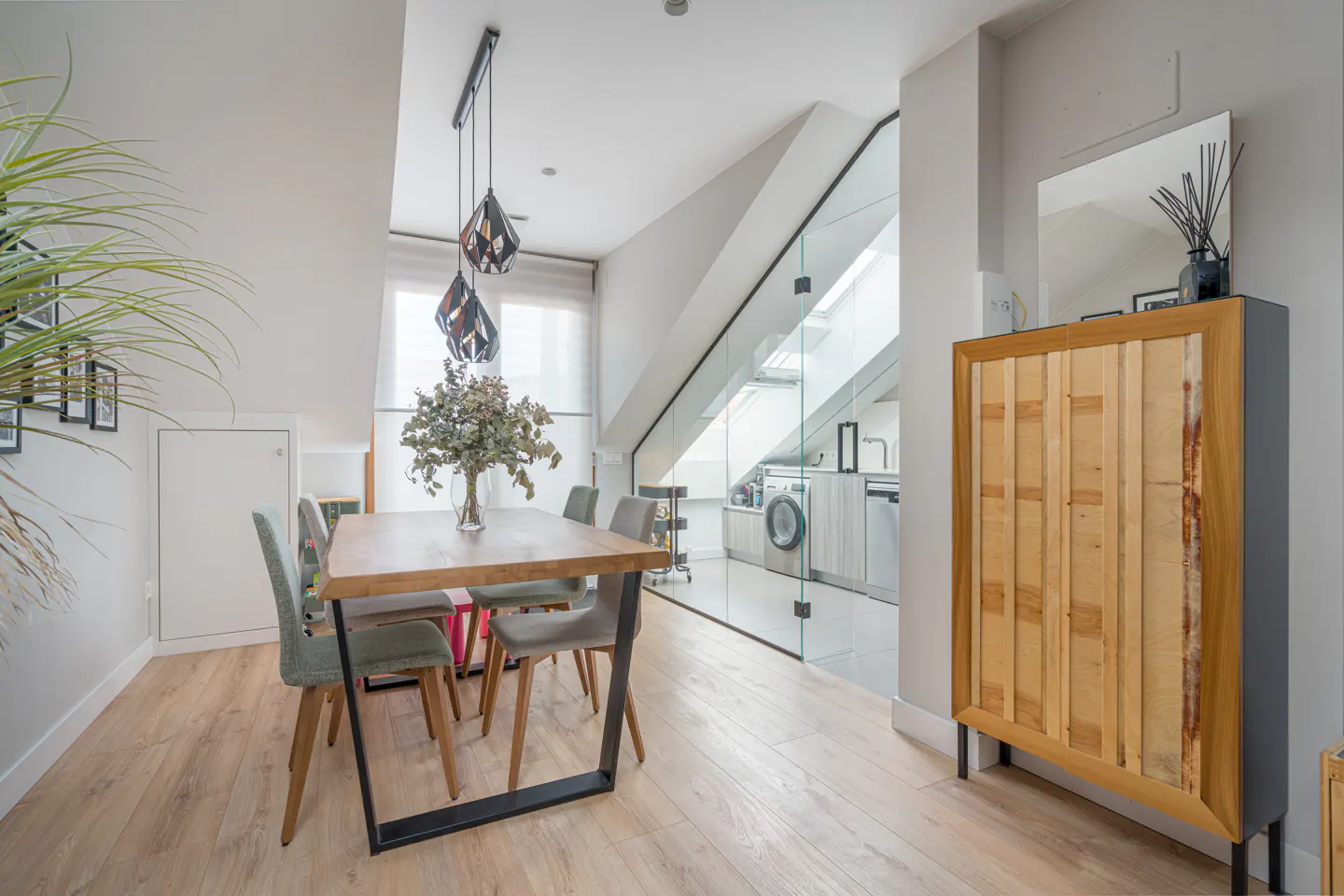 Bright dining area with wood table, gray chairs, and geometric pendant lights. Glass wall reveals a laundry area. Cabinet with mirror stands to the right.