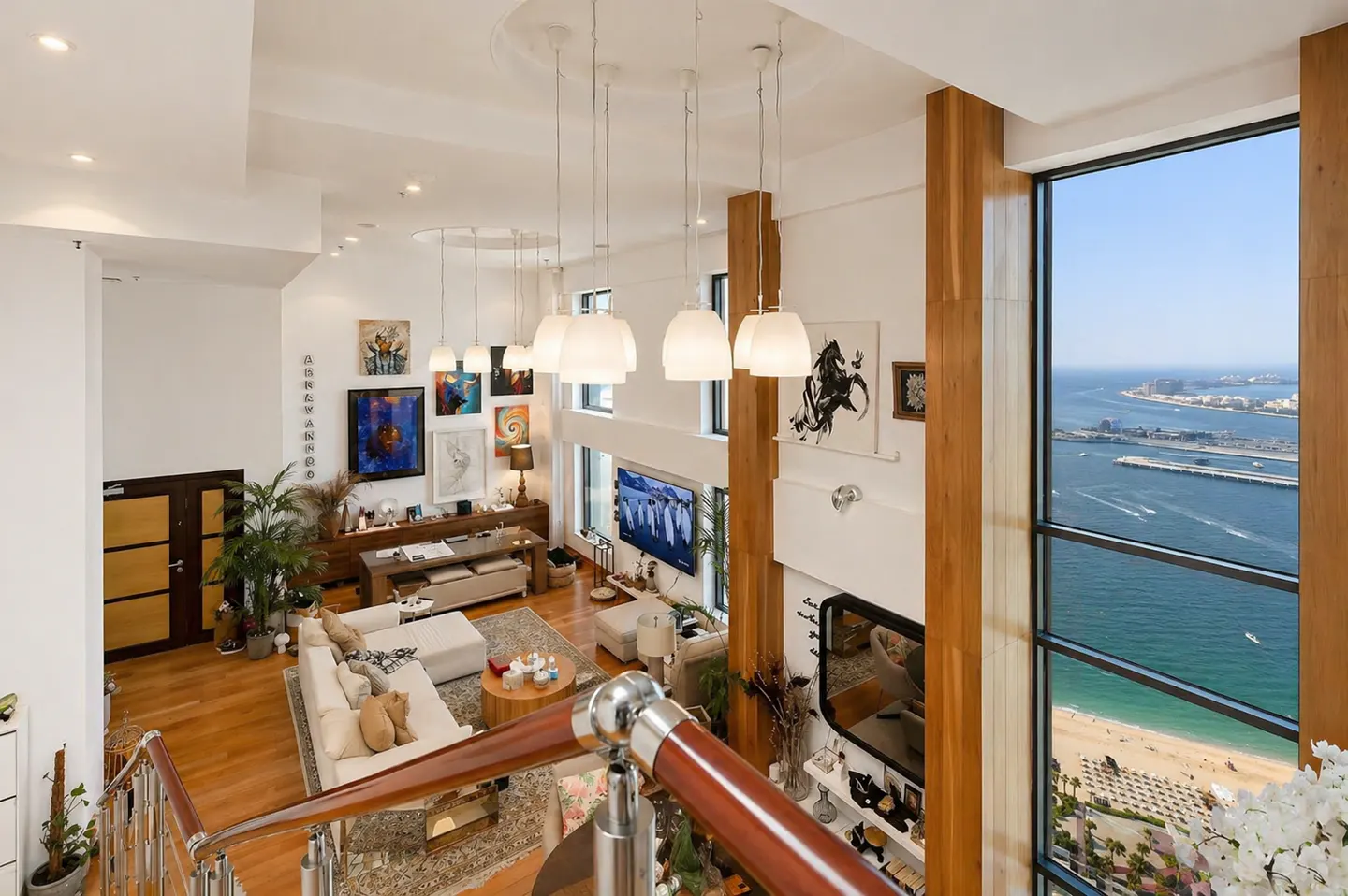 A high-angle view of a modern living room with a large window overlooking a beach and ocean. The room has white walls, wood floors, and pendant lights.