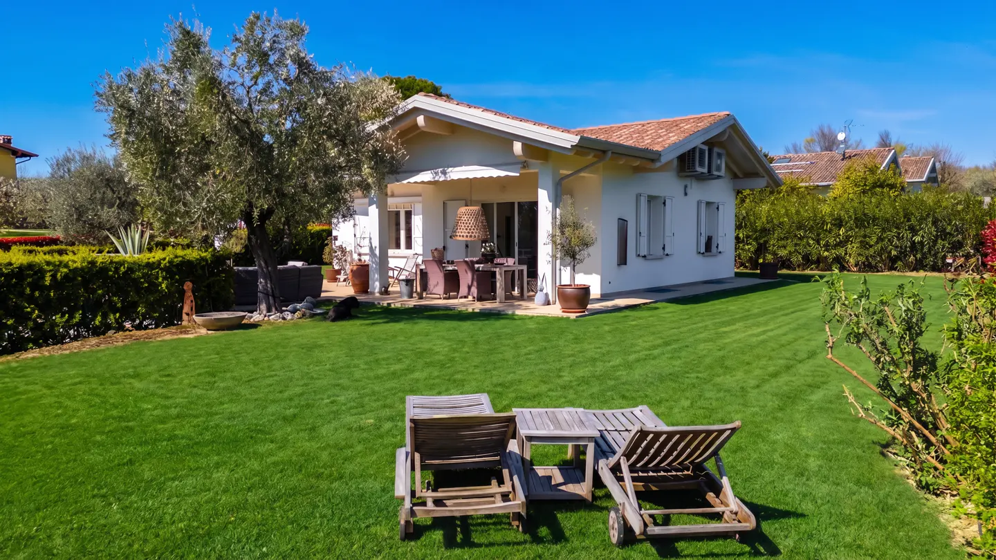 Exterior view of a white house with a red tile roof, green lawn, and patio furniture under a blue sky.