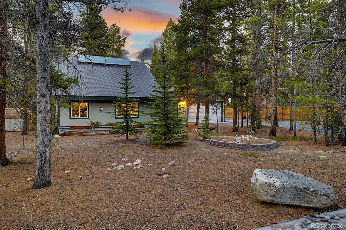 A cozy, light-green cabin with a metal roof sits nestled among tall pine trees at dusk. Solar panels are visible on the roof.