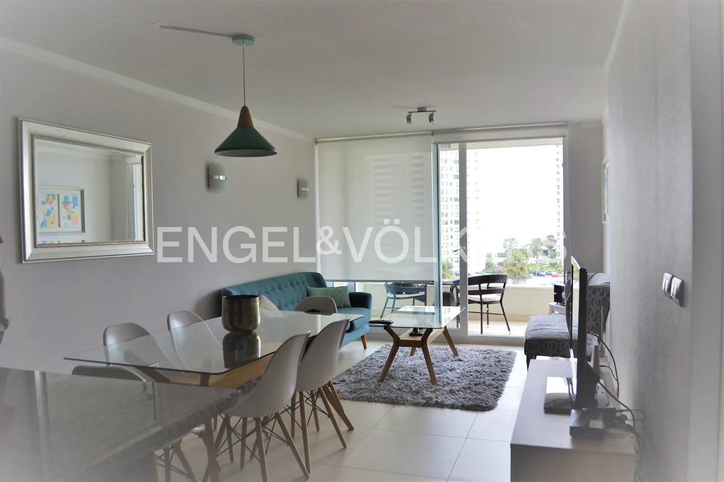 Bright living room with a glass table, white chairs, a blue sofa, and a balcony with a view.