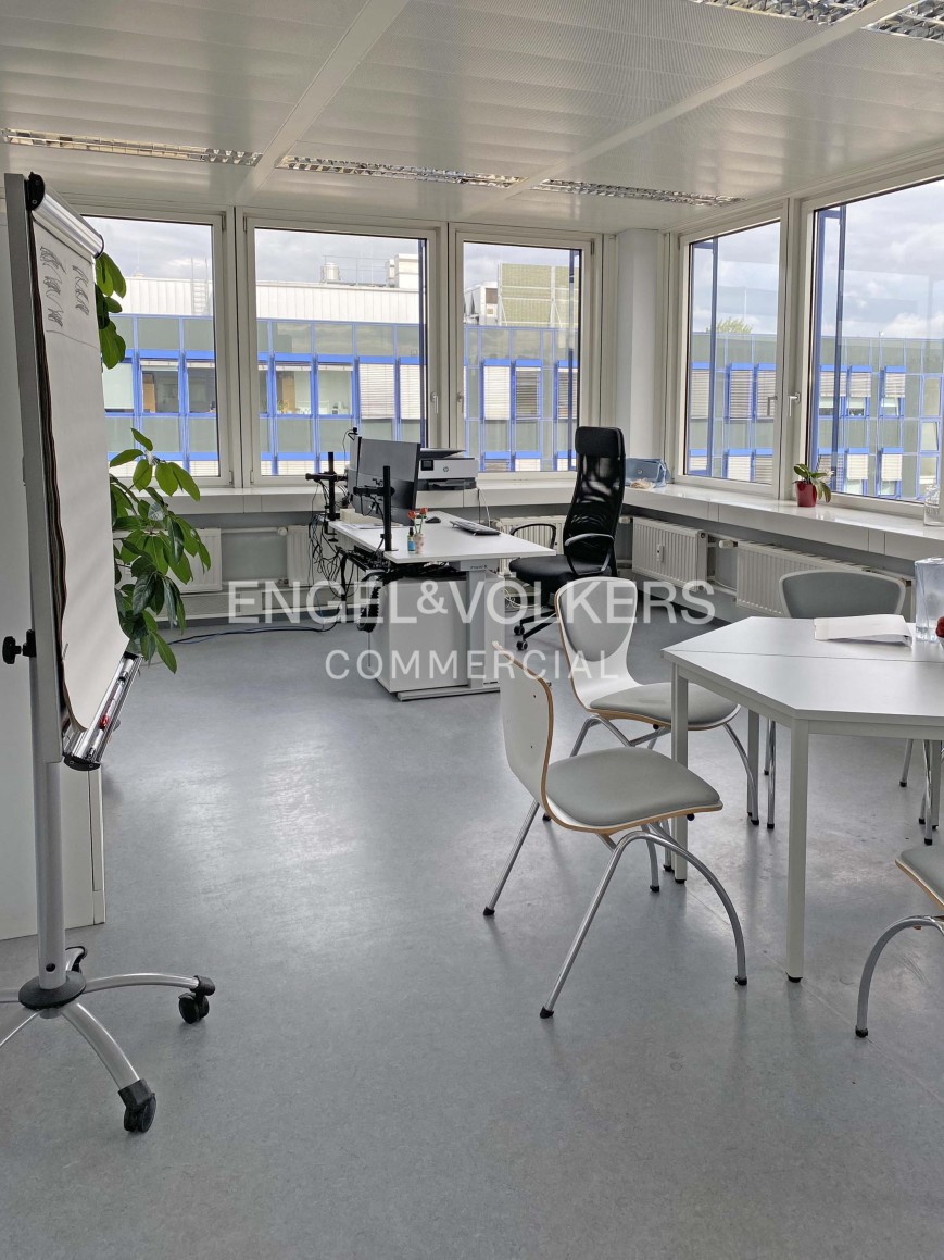 Modern office space with white desks, chairs, a whiteboard on wheels, and large windows overlooking a building exterior. [AI-generated text]
