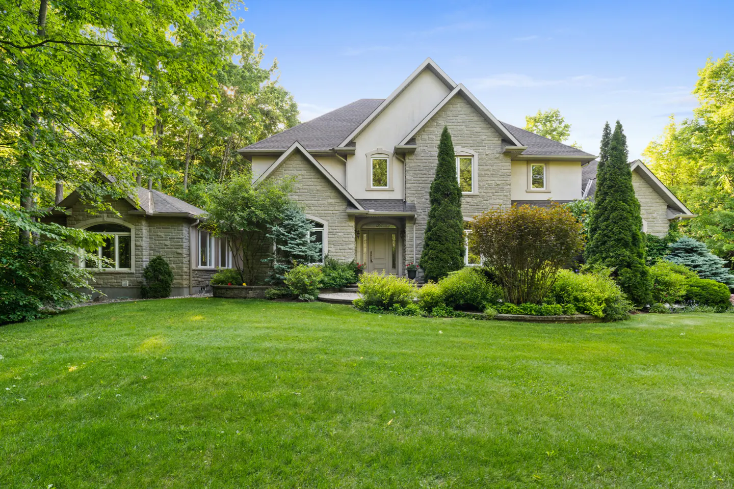 Large stone and stucco house with a green lawn, surrounded by lush trees and manicured landscaping under a blue sky.