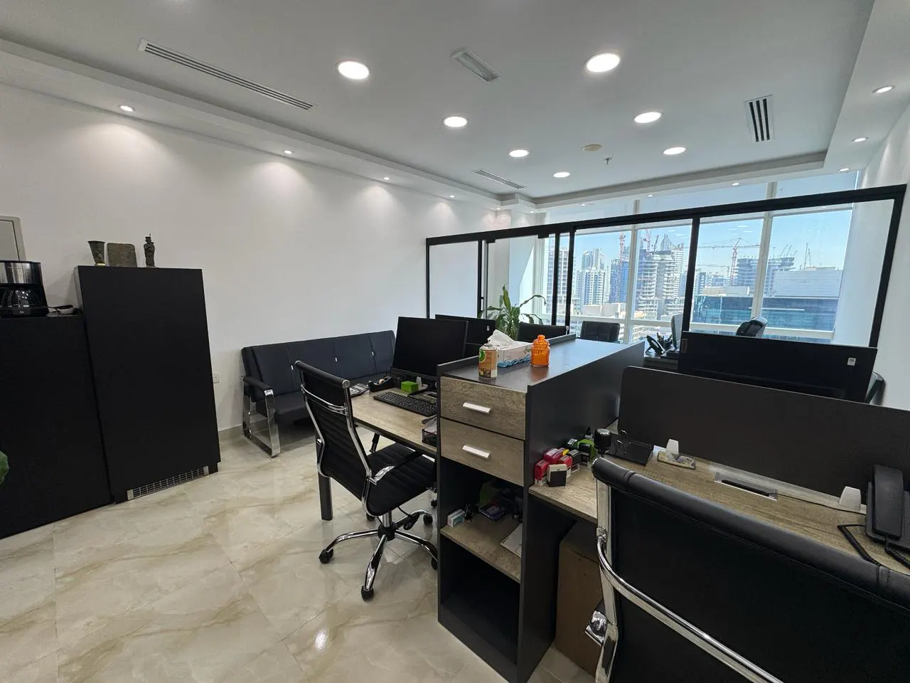 Modern office with black desks, ergonomic chairs, and large windows overlooking a city skyline with construction cranes.