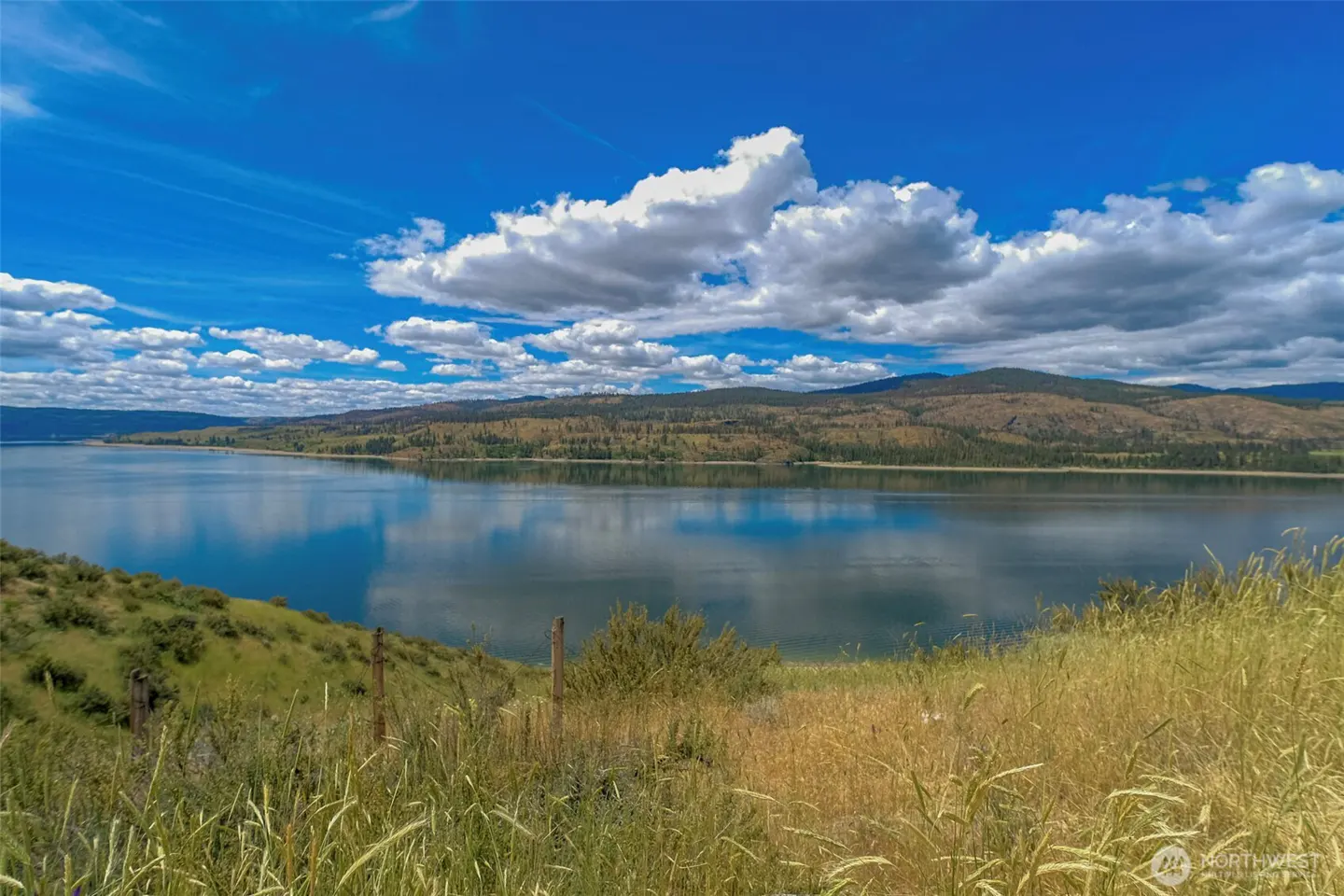 A scenic view of a calm lake under a bright blue sky with fluffy white clouds, framed by grassy hills in the foreground.