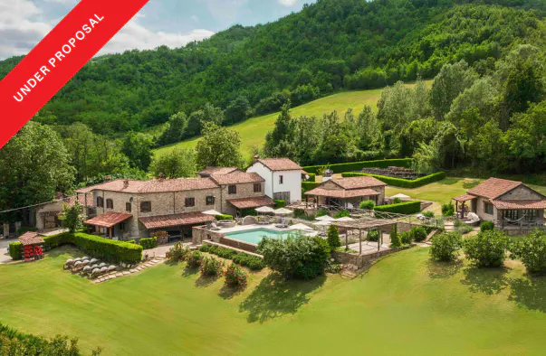 Aerial view of a stone house with a pool, surrounded by green hills and trees. "Under Proposal" banner in the upper left corner.