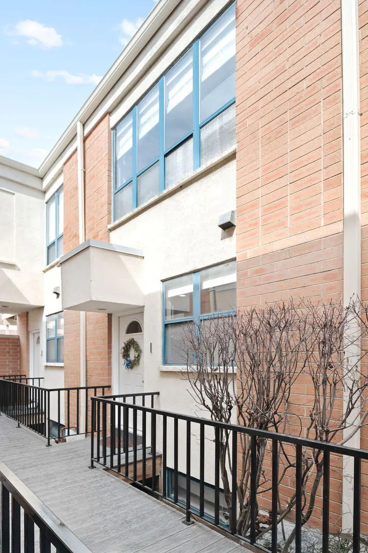 Exterior view of a townhouse with a brick facade, blue-framed windows, and a white door with a wreath. A wooden walkway with black railings leads to the entrance.