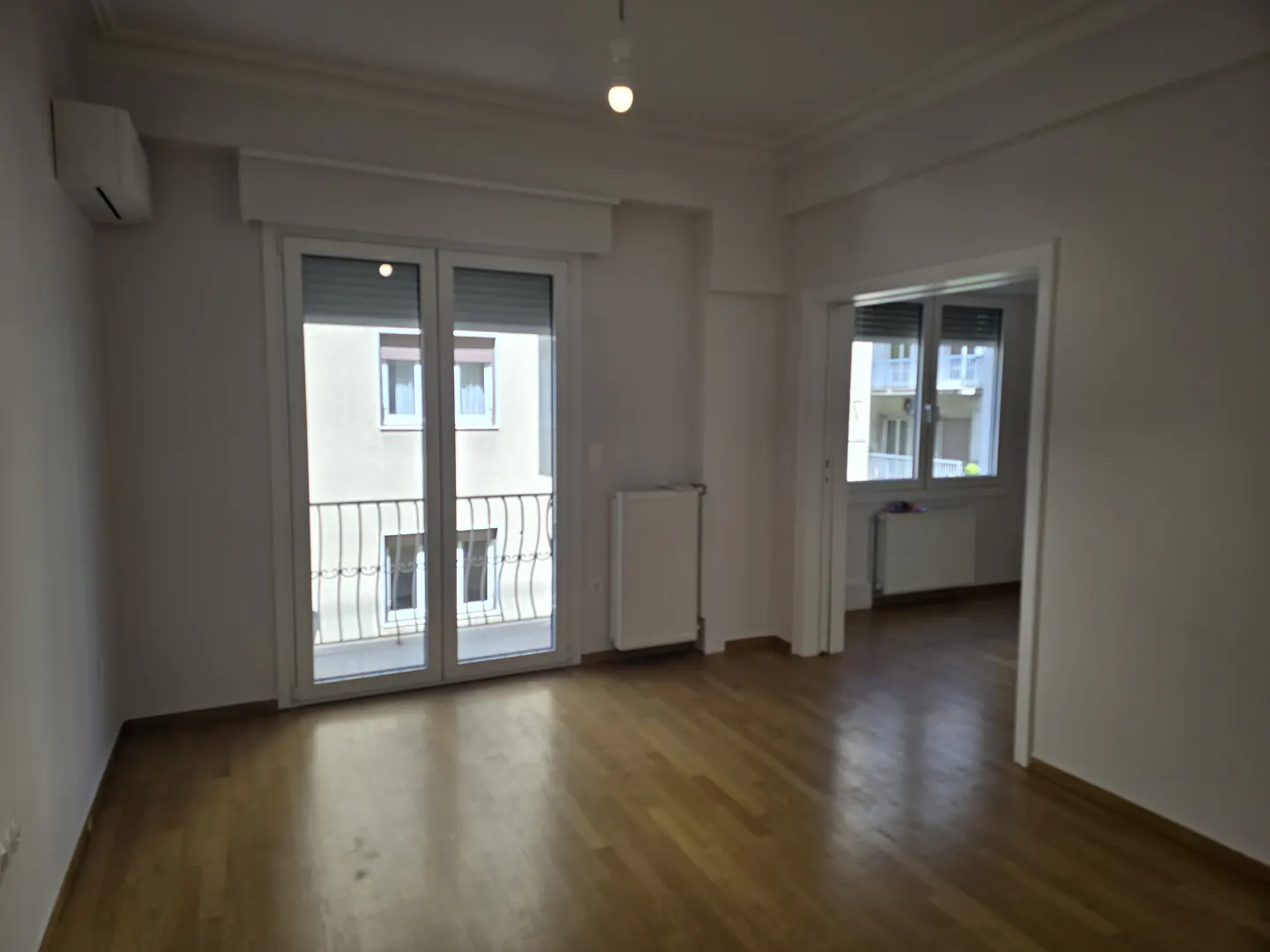 Bright, empty room with hardwood floors, white walls, and a balcony with a view of a building across the street.