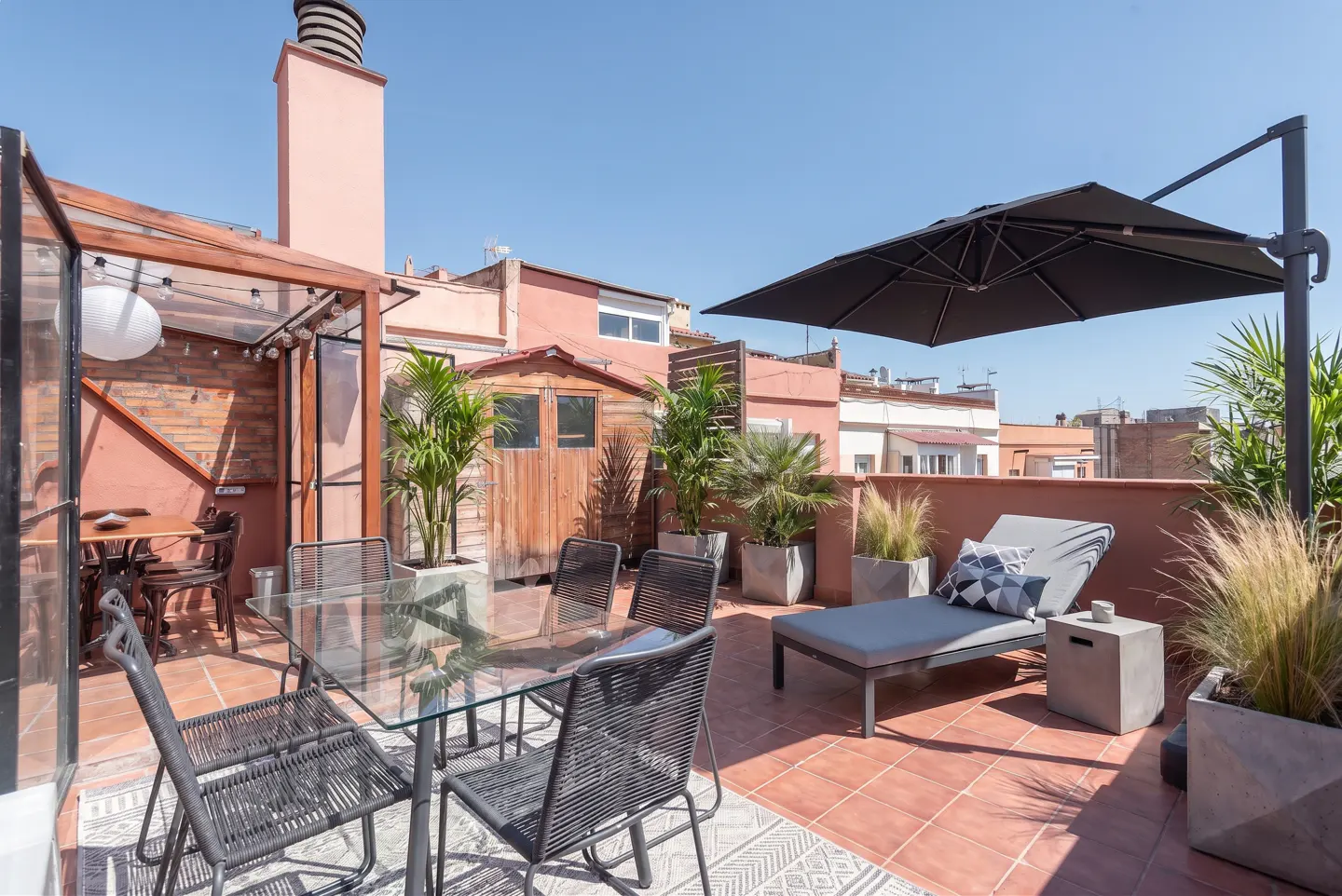 Rooftop patio with a glass table, black chairs, a gray lounge chair under a black umbrella, and potted plants.