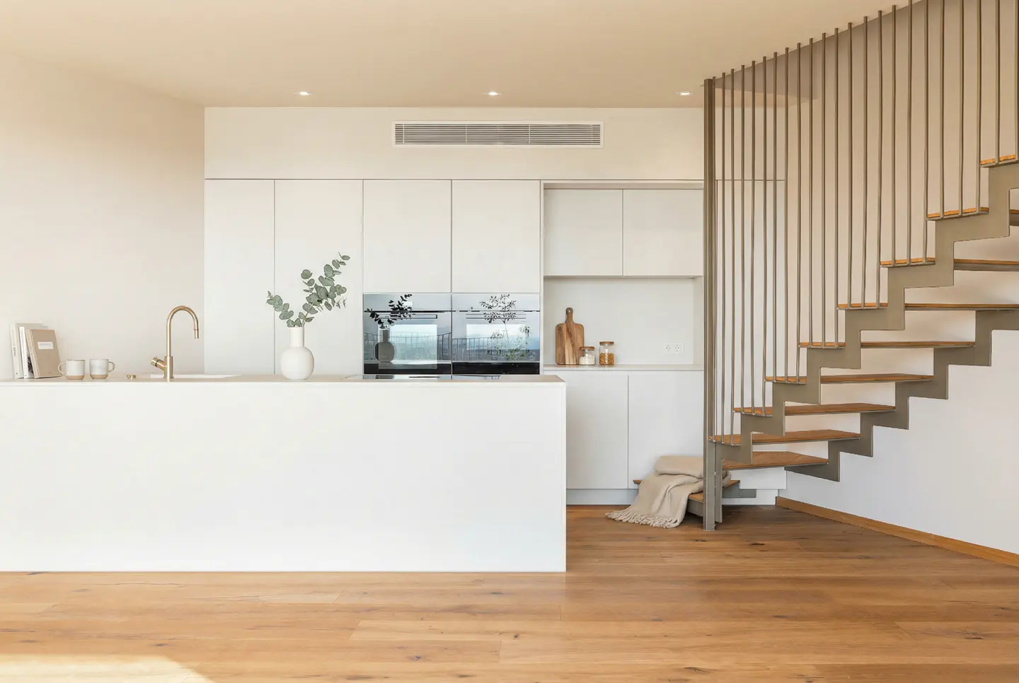 Bright, modern kitchen with white cabinets, island, and wood floors. Stairs with metal railings are on the right. A vase with greenery sits on the island.
