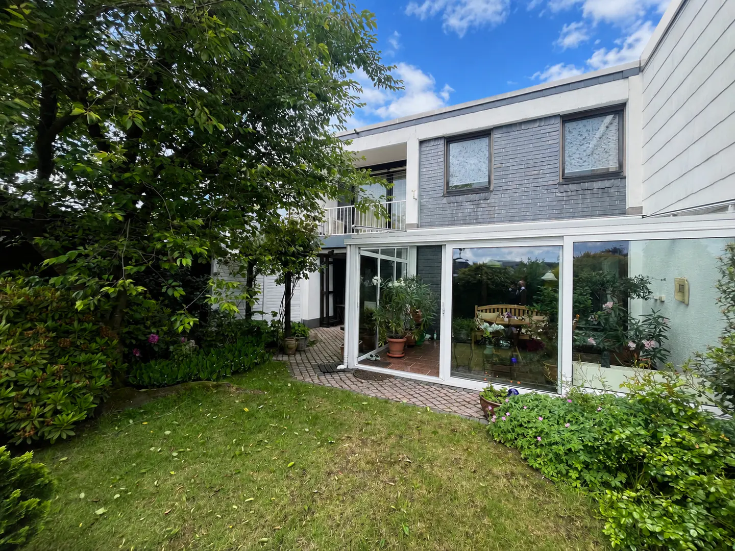 Backyard view of a two-story house with a glass-enclosed sunroom, brick patio, lush green lawn, and large shade tree.