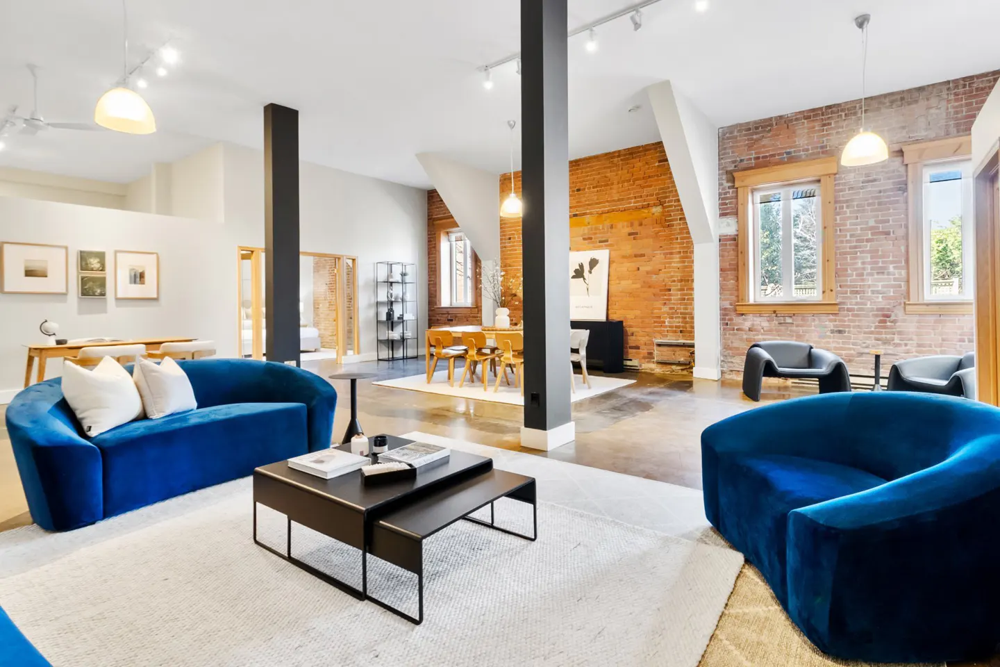 Bright, open-concept living space with blue velvet sofas, black coffee tables, and exposed brick walls. Dining area visible in the background.