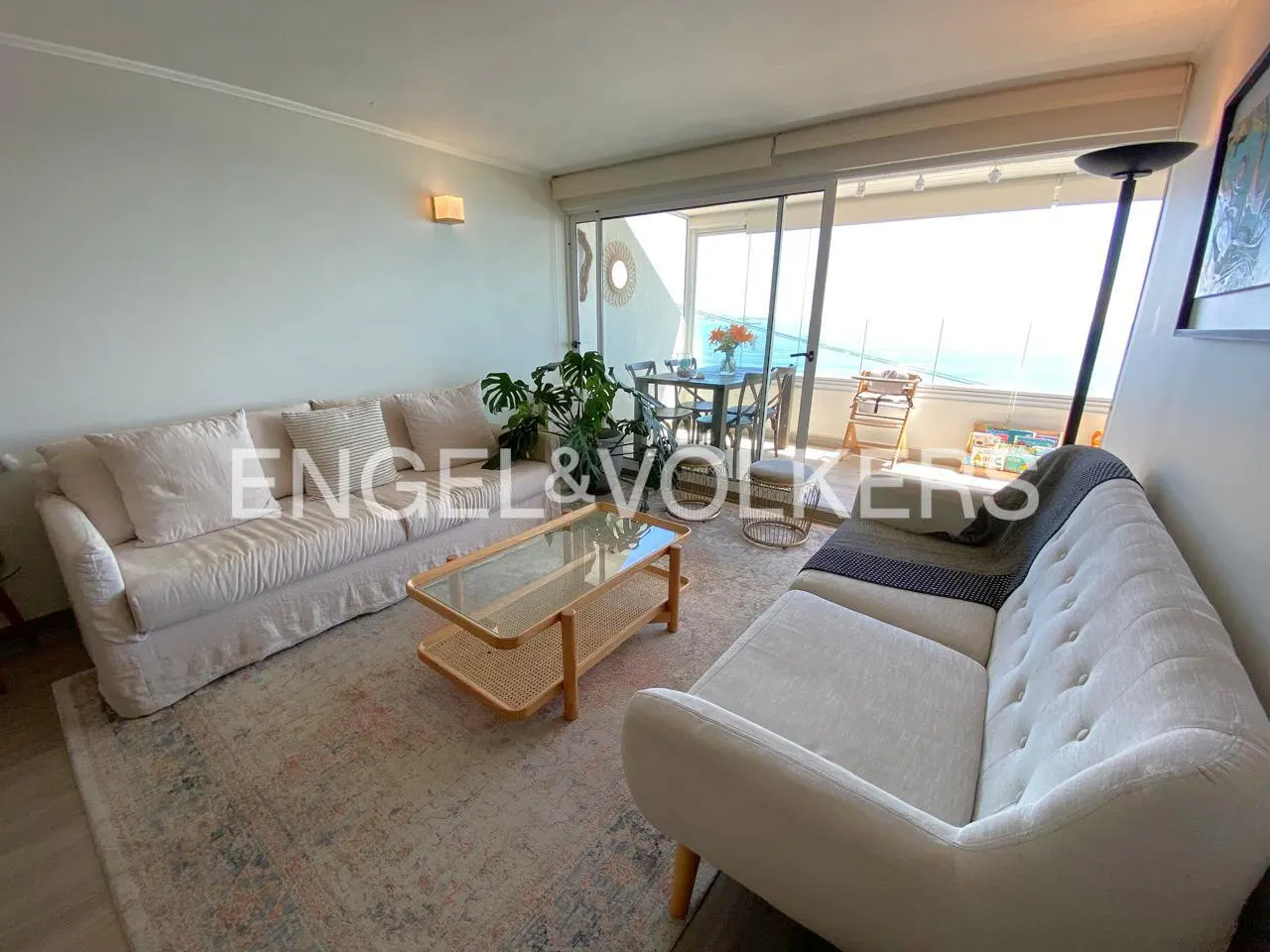 Living room with two white sofas, a glass coffee table, and a dining area with ocean views. Engel & Völkers logo visible.