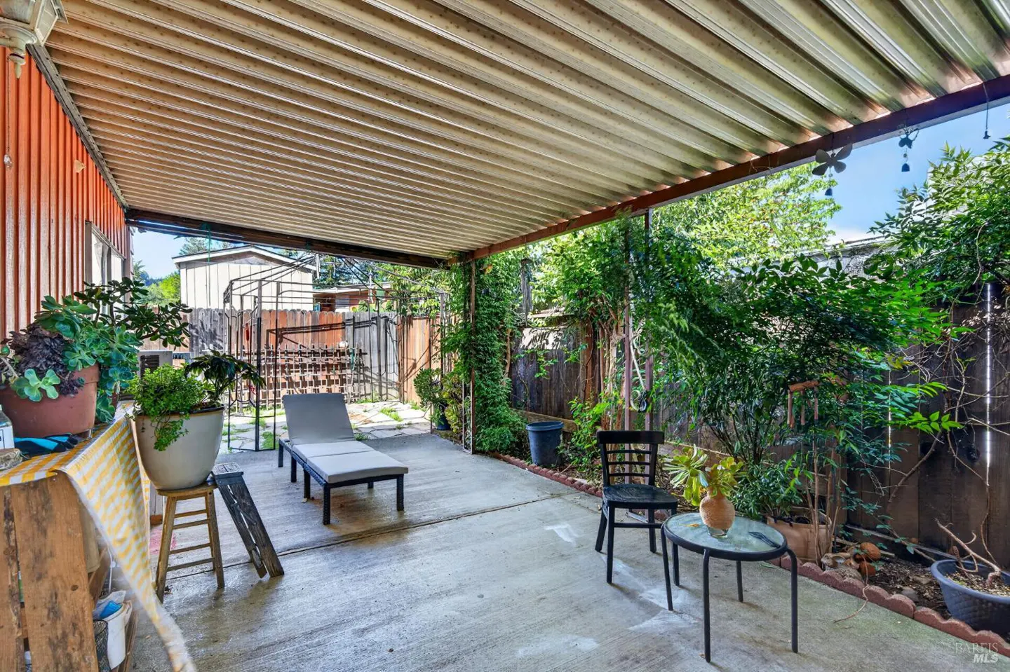 Covered patio with a lounge chair, table, and chair. Green plants line the fence.
