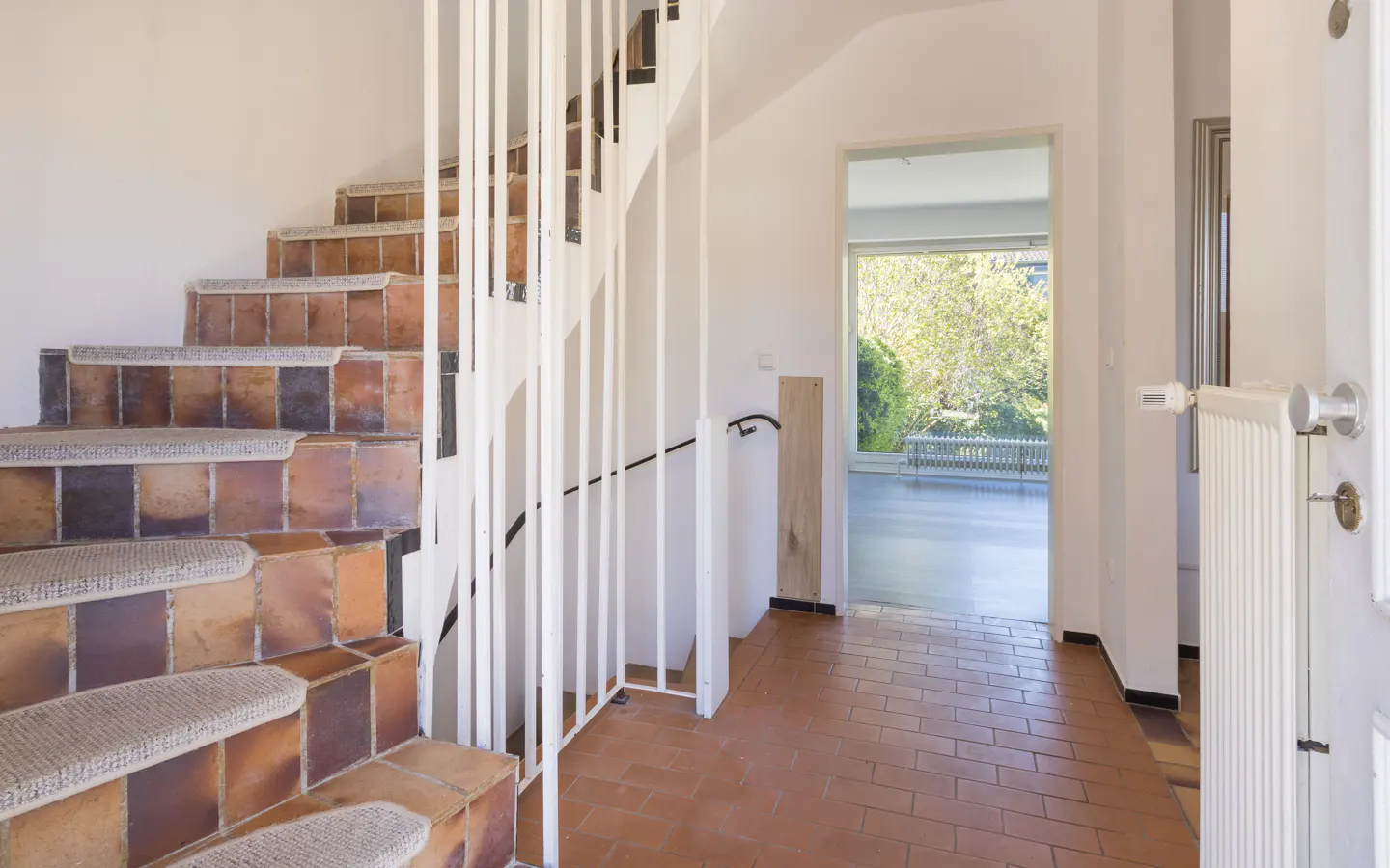 Interior view of a home featuring tile stairs with carpet treads and a white railing, leading to a doorway with a view of greenery.