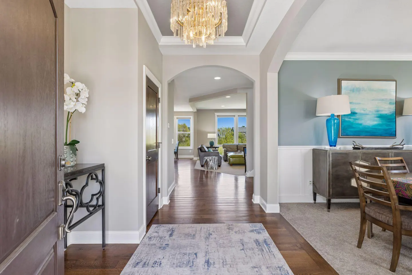 Open front door reveals a foyer with hardwood floors, a crystal chandelier, and a view into the living room.