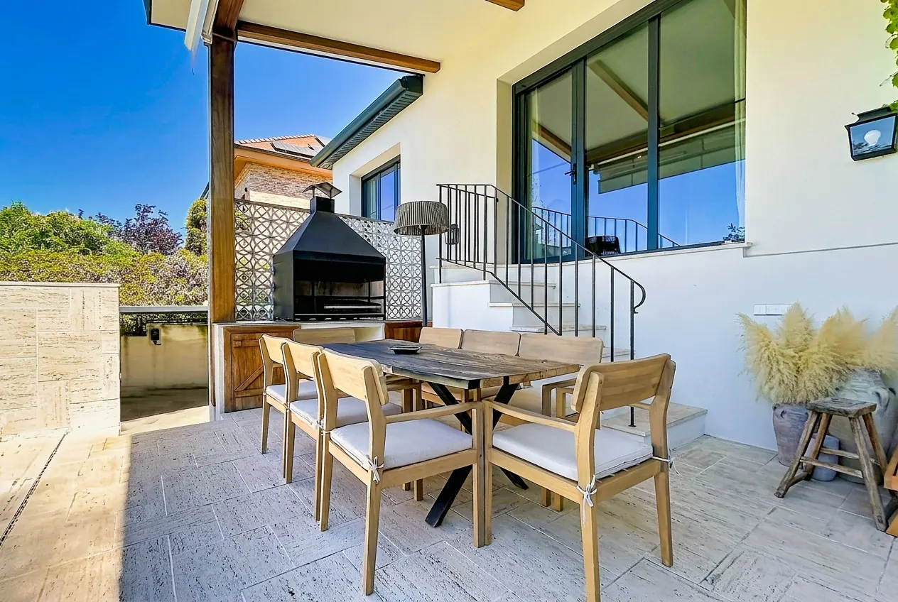 Outdoor patio with a wooden table and chairs, a black grill, and stairs leading to a white building.