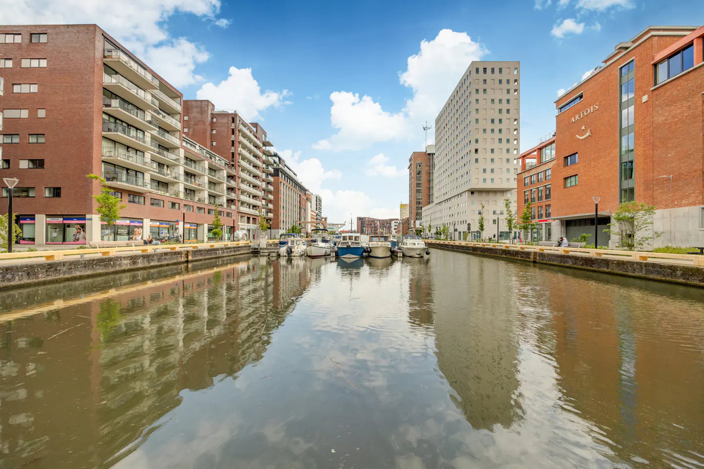 A canal lined with modern brick apartment buildings and boats, reflecting the blue sky and clouds on the water's surface.