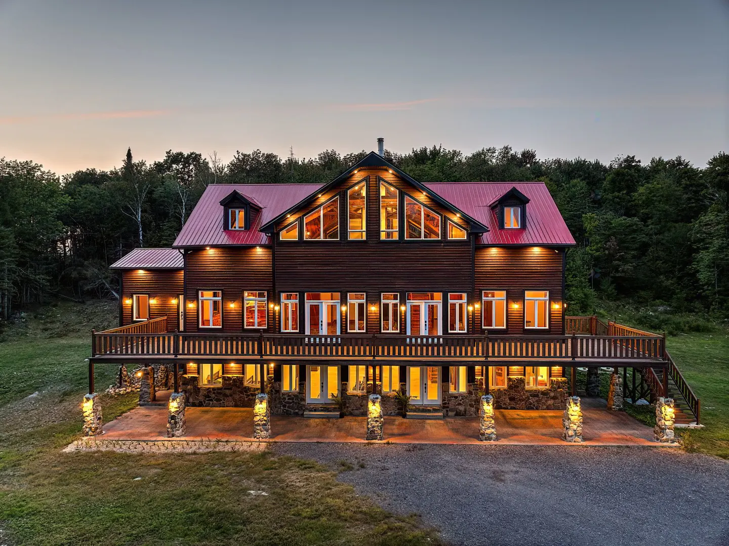 A large, two-story brown house with a red roof and a wide porch, set against a backdrop of trees and a dusky sky.