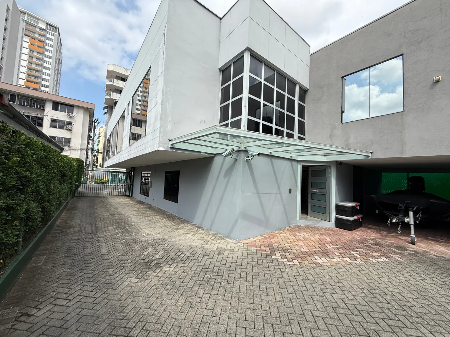 Exterior of a modern gray building with large windows and a glass awning over the entrance. A brick driveway leads to a gated entrance.