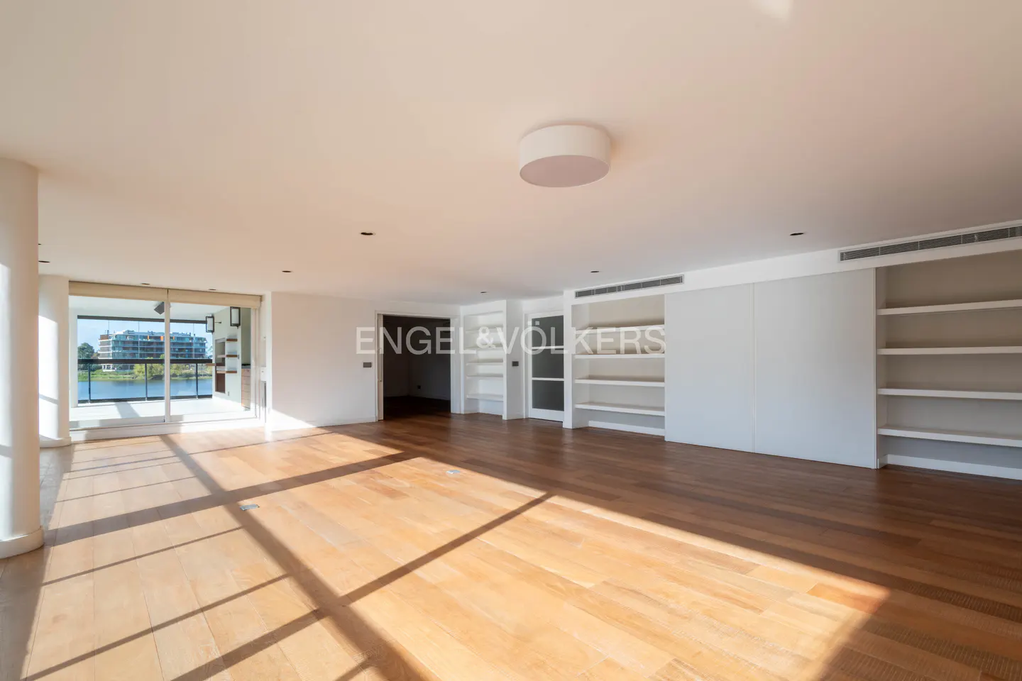 Bright, empty room with hardwood floors and white walls. A large window overlooks a lake. Built-in shelves line one wall.