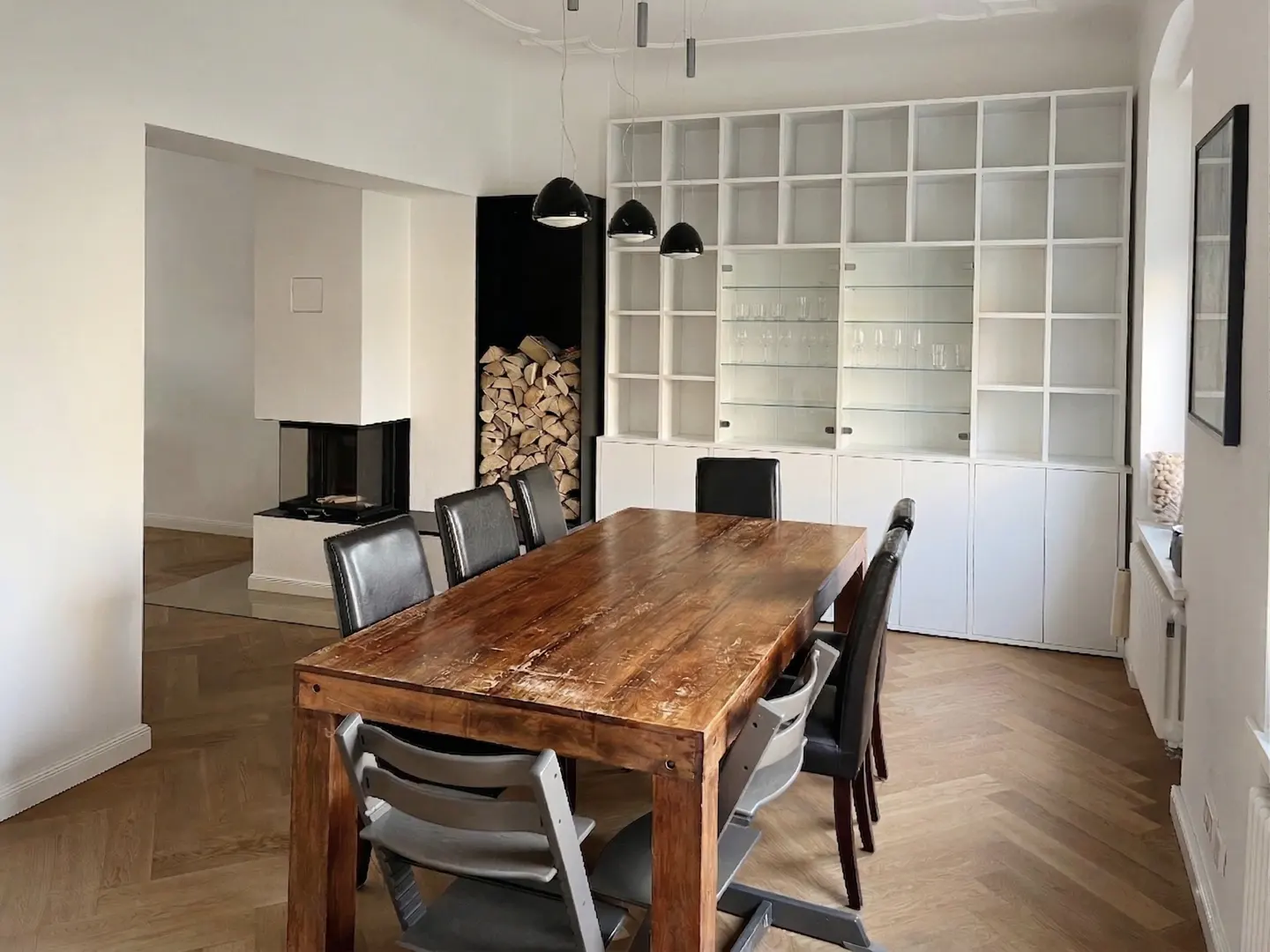 A dining room with a wooden table, black chairs, a white fireplace, and a white bookshelf.