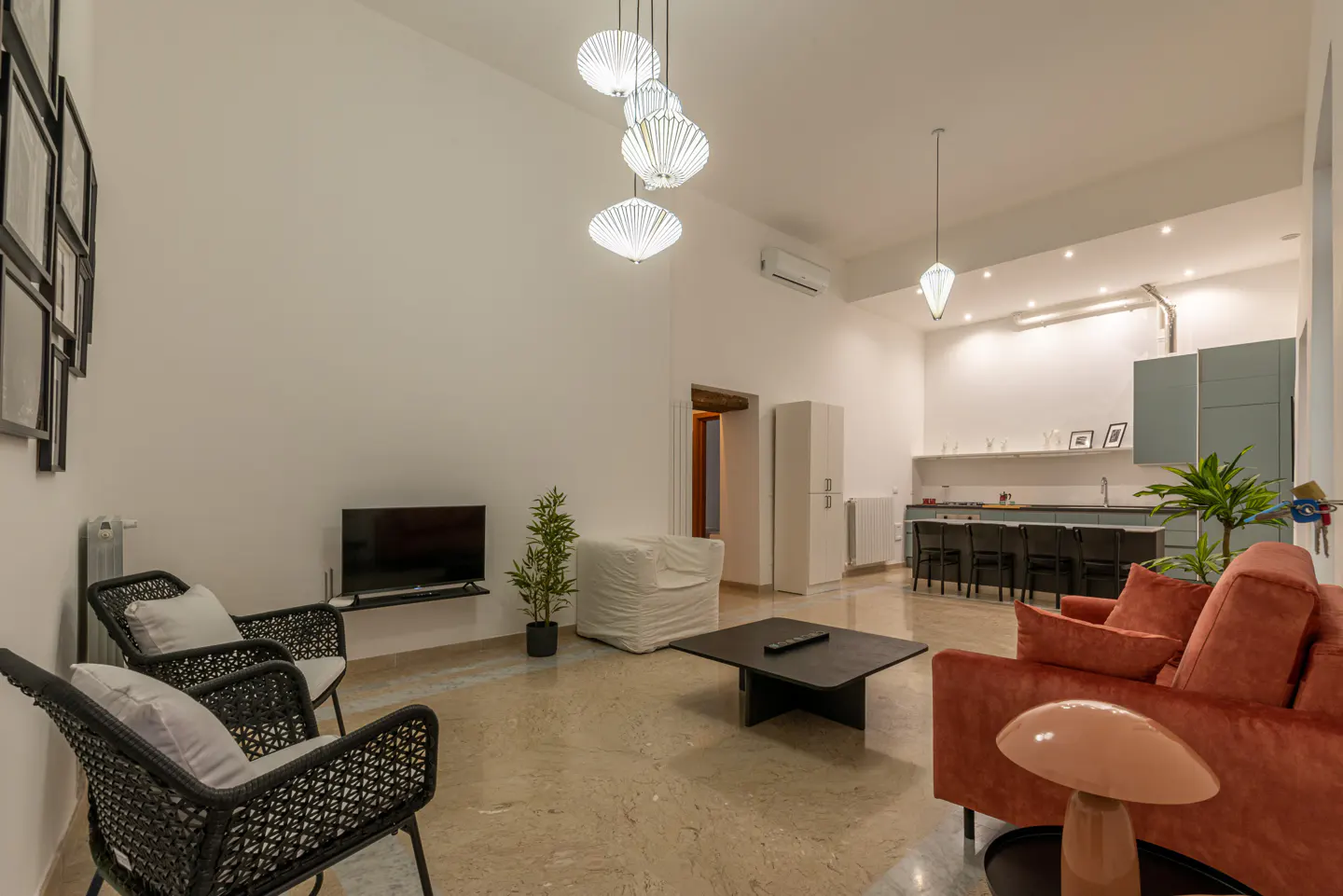 Bright, open living space with a coral sofa, black wicker chairs, and a modern kitchen in the background. White walls and unique lighting fixtures.