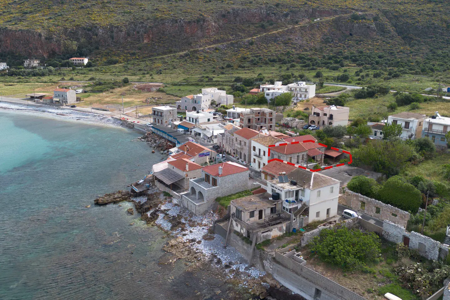 Aerial view of coastal village with stone buildings, red tile roofs, and turquoise water. Mountain backdrop.