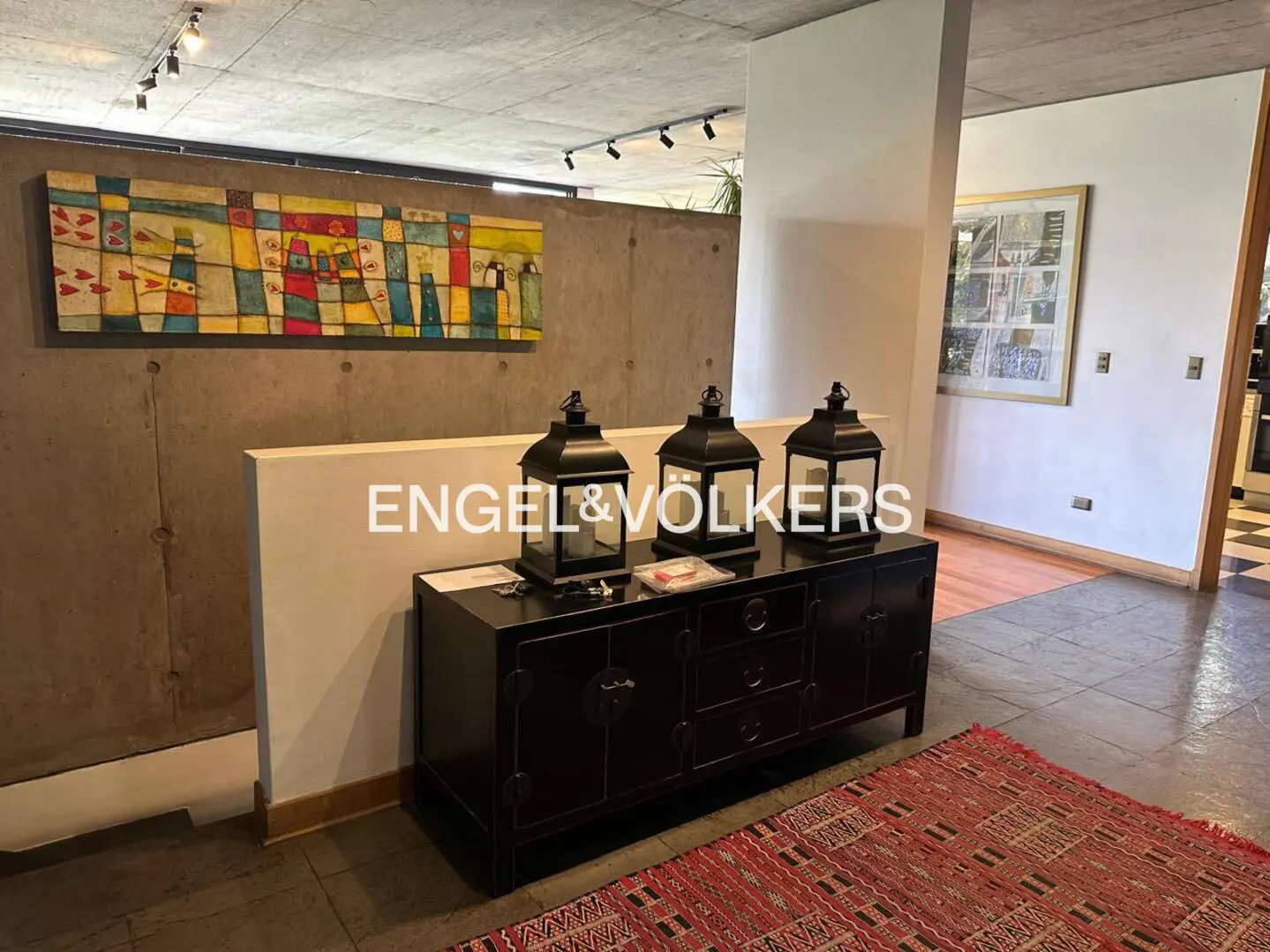Interior view of a modern home with a black cabinet topped with three lanterns, a colorful painting, and a red rug.