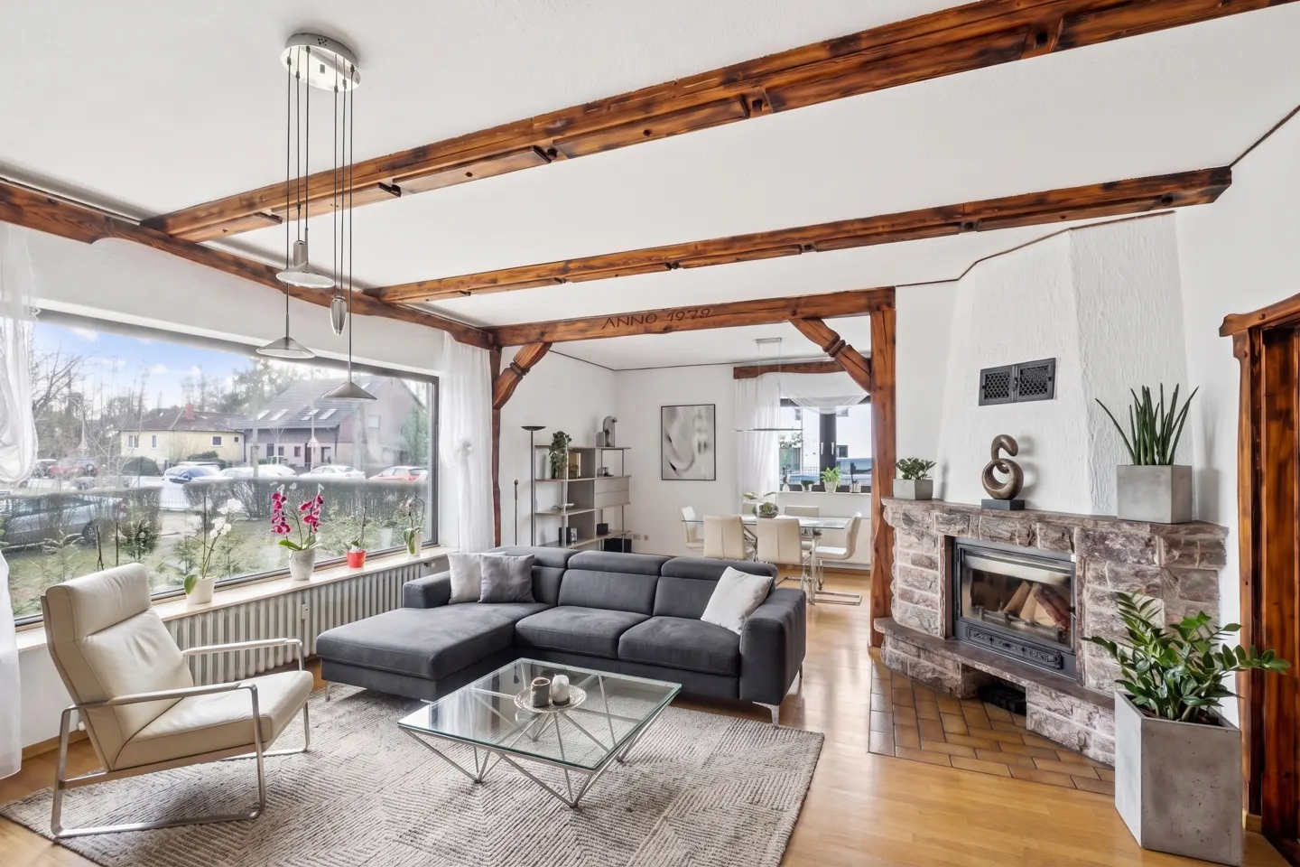 Living room with a gray sectional sofa, glass coffee table, stone fireplace, and wooden beams. A large window overlooks a canal.