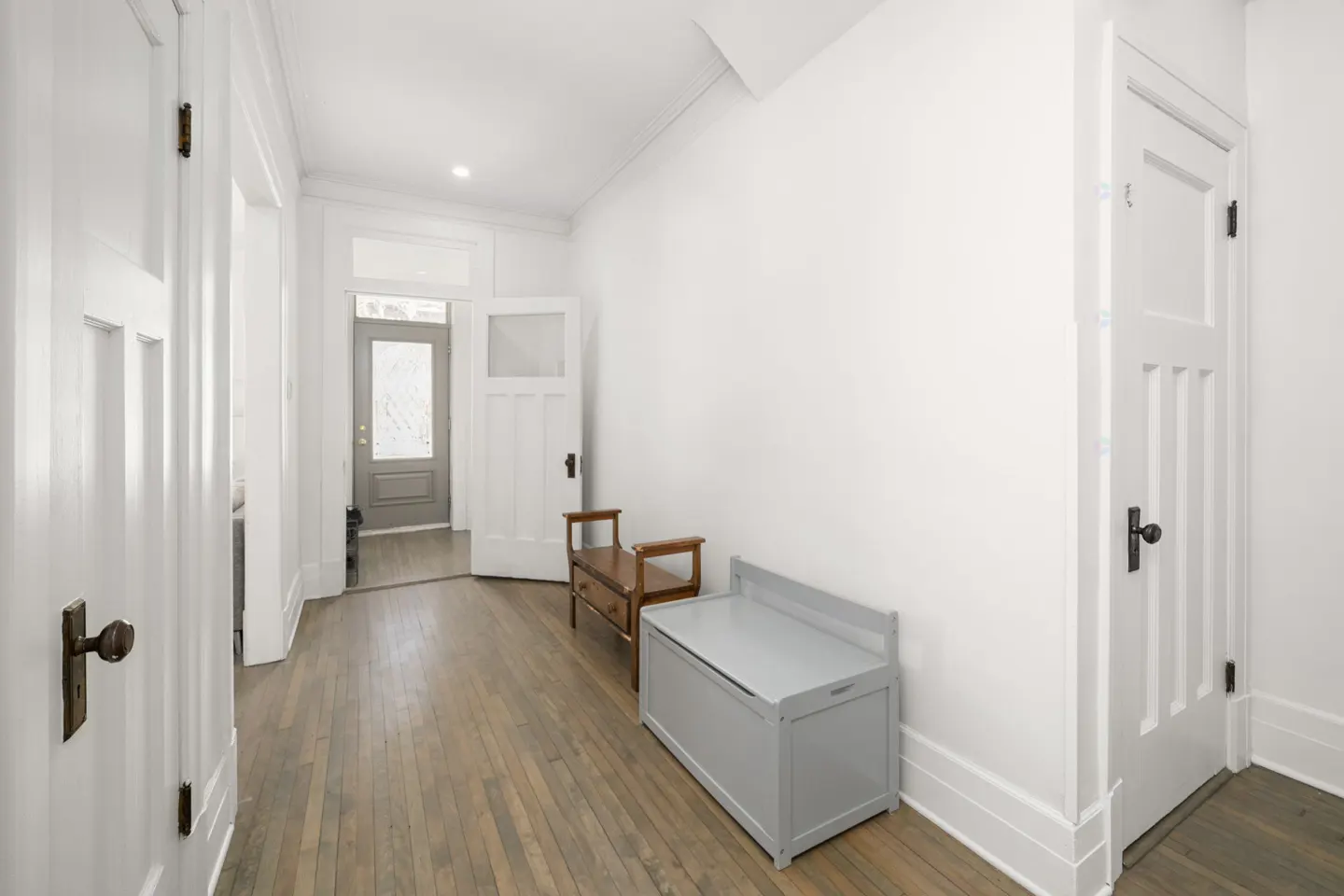 Bright hallway with hardwood floors, white walls, and white doors. A grey storage bench and wooden bench sit against the wall. An open door leads to the outside.