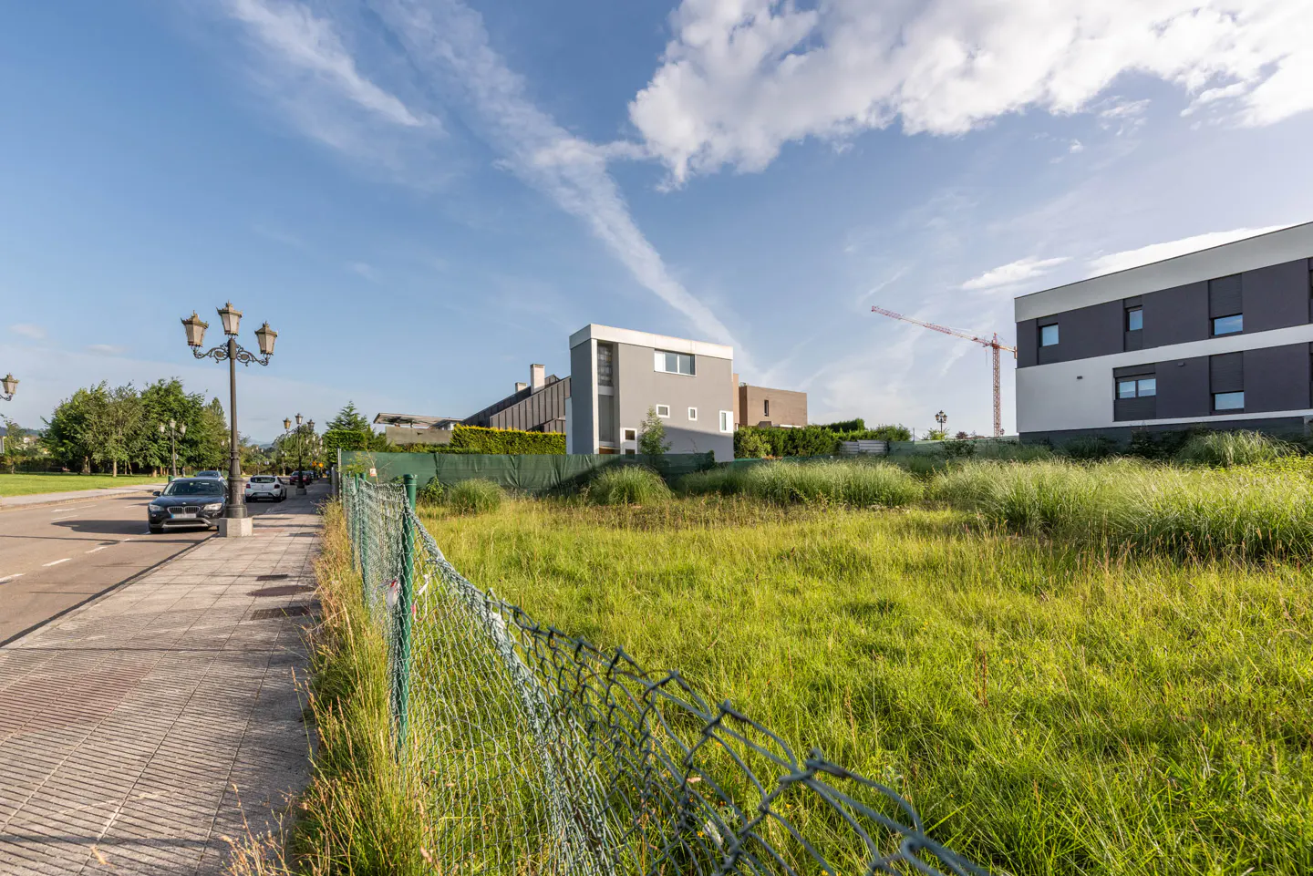 A grassy vacant lot enclosed by a wire fence next to a sidewalk, with modern apartment buildings under a blue sky.