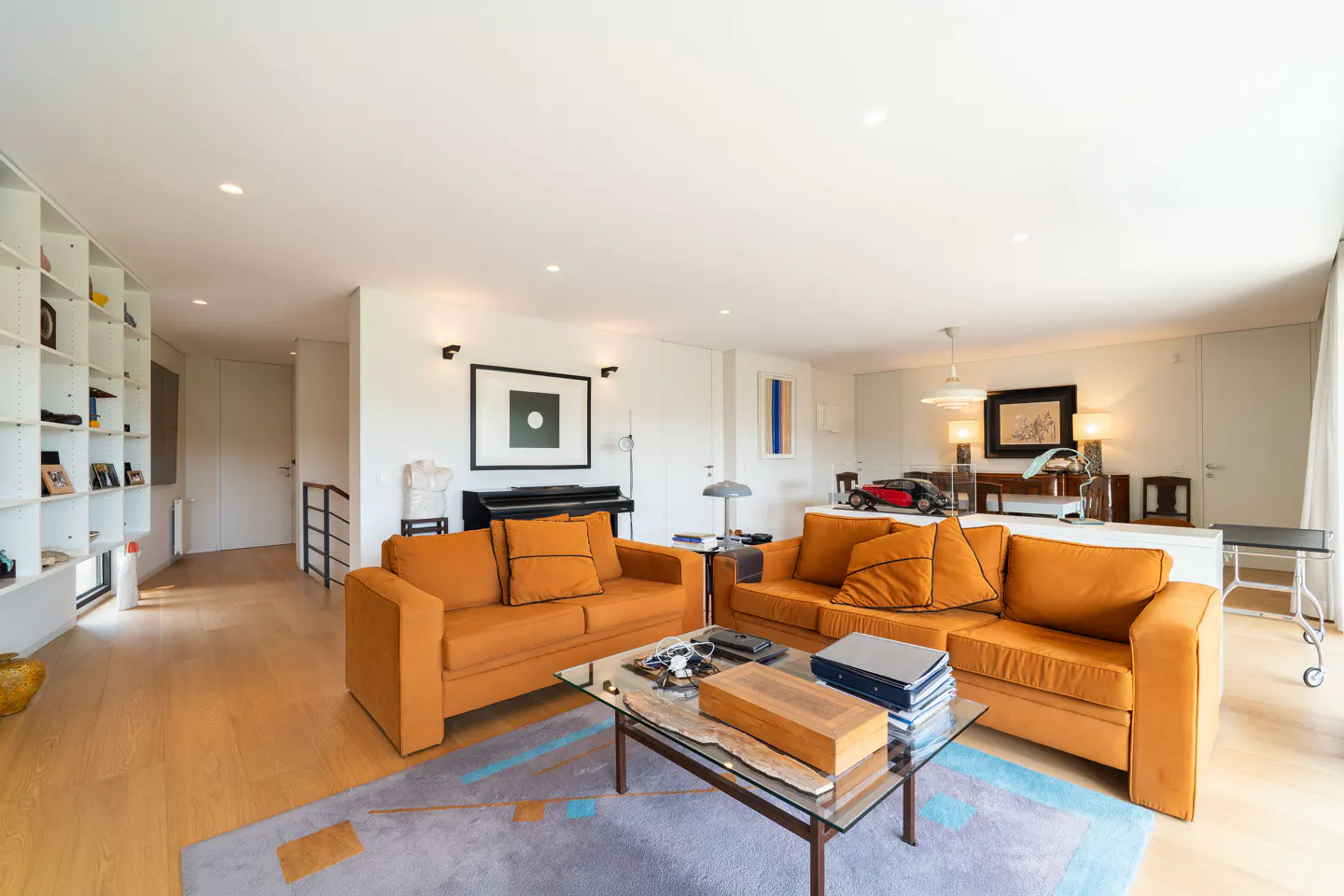 Bright living room with hardwood floors, orange sofas, and a glass coffee table on a blue rug. A piano and white shelves line the back wall.