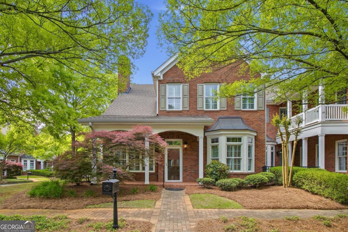 Two-story red brick house with a front porch, bay window, and a brick walkway surrounded by lush green trees and shrubs.
