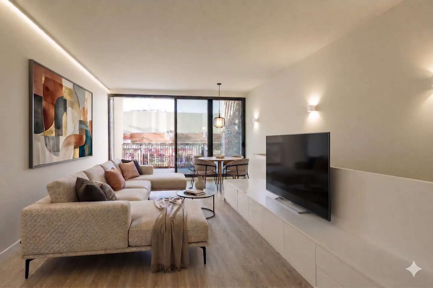Bright living room with beige sectional sofa, abstract art, and a TV on a white console. A round table and chairs are near a sliding glass door.