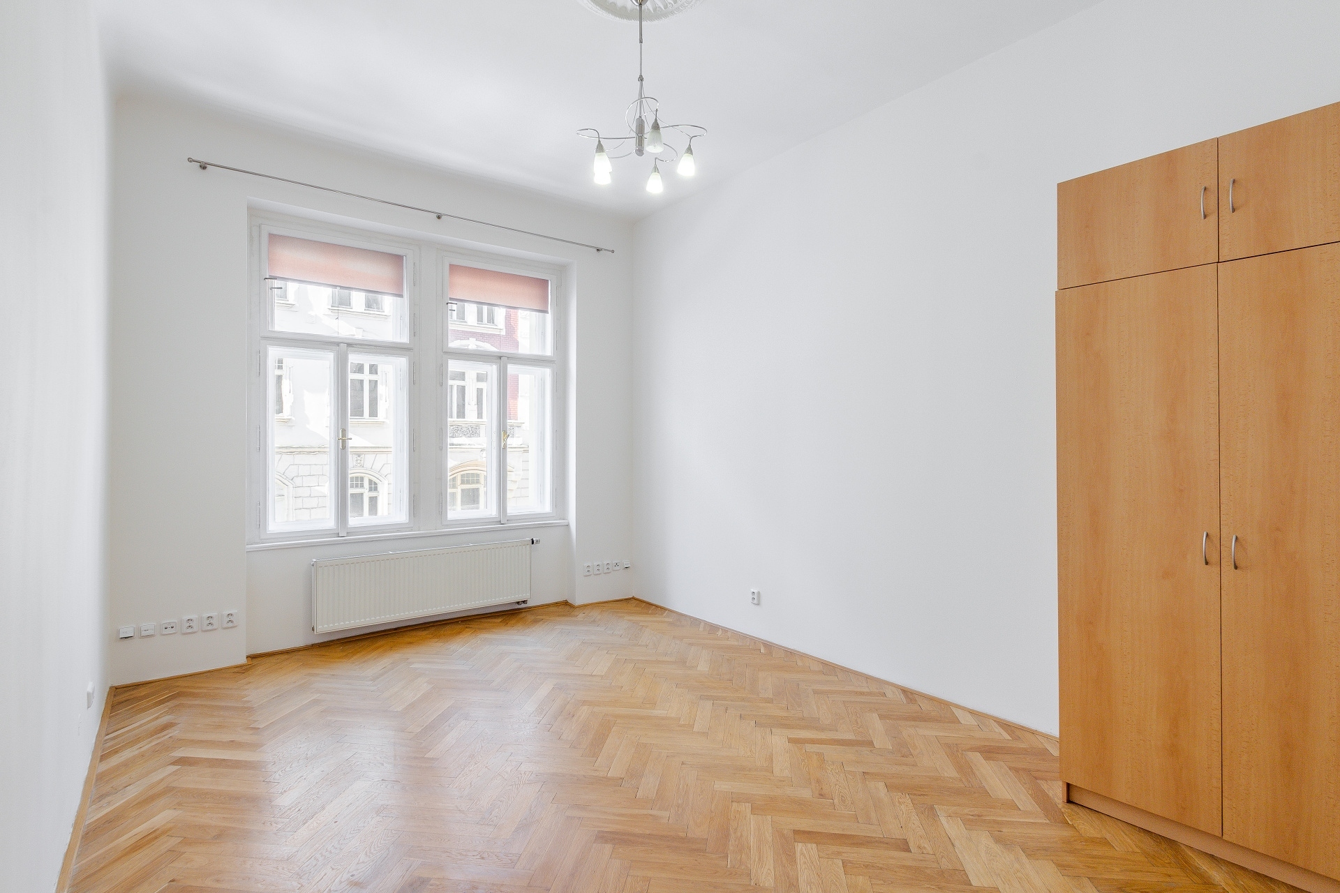 Empty room with white walls, herringbone wood floors, a large window, a modern chandelier, and a tall light wood wardrobe. [AI-generated text]