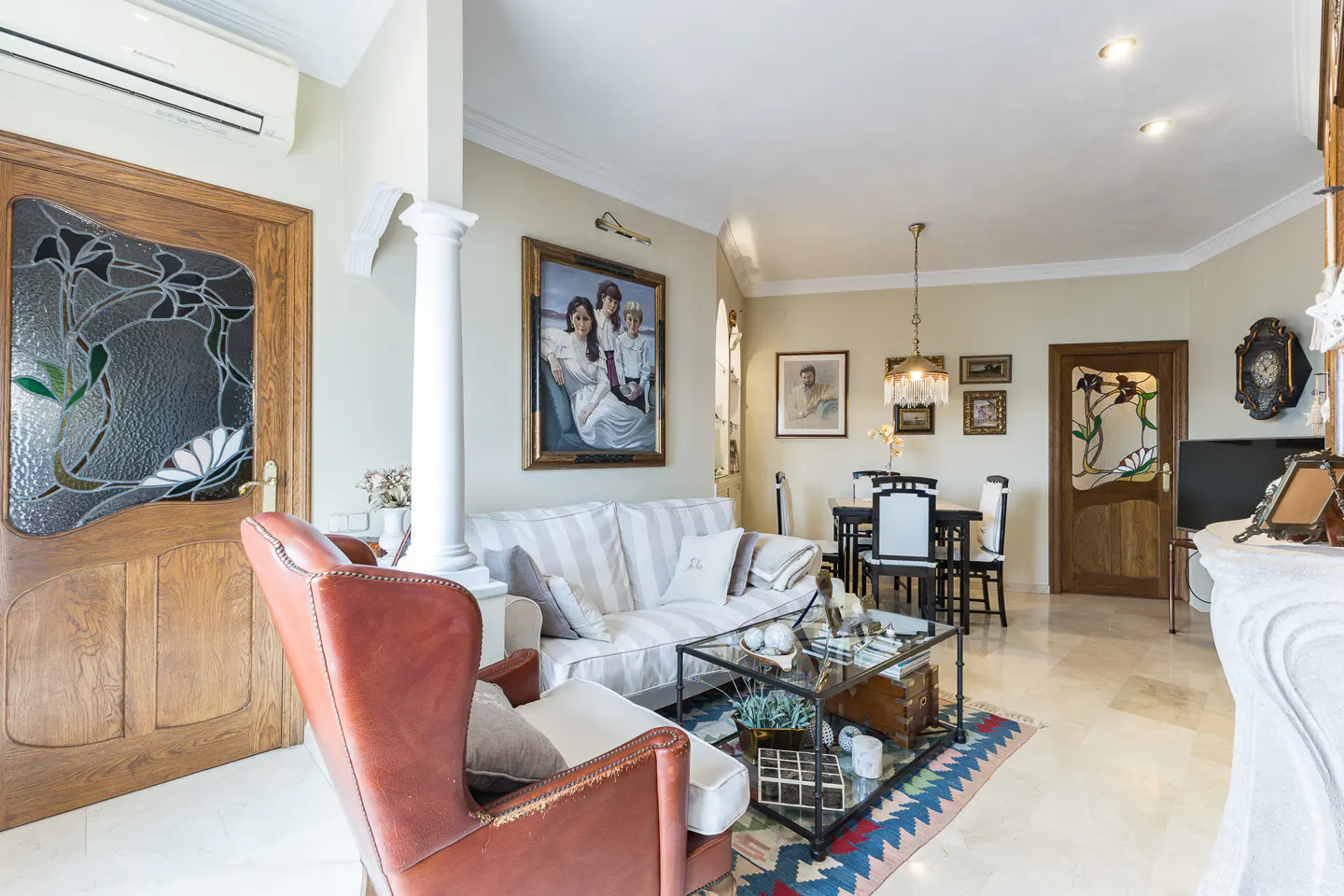 A bright living room with a brown leather chair, striped sofa, glass coffee table, and dining table. Art Nouveau doors and paintings decorate the walls.