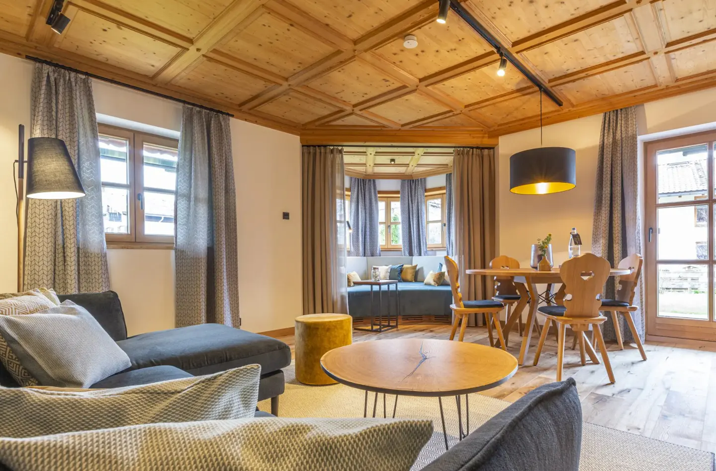 A cozy living room with wood ceiling, gray sofa, round wood table, and dining set. Natural light streams through curtained windows.