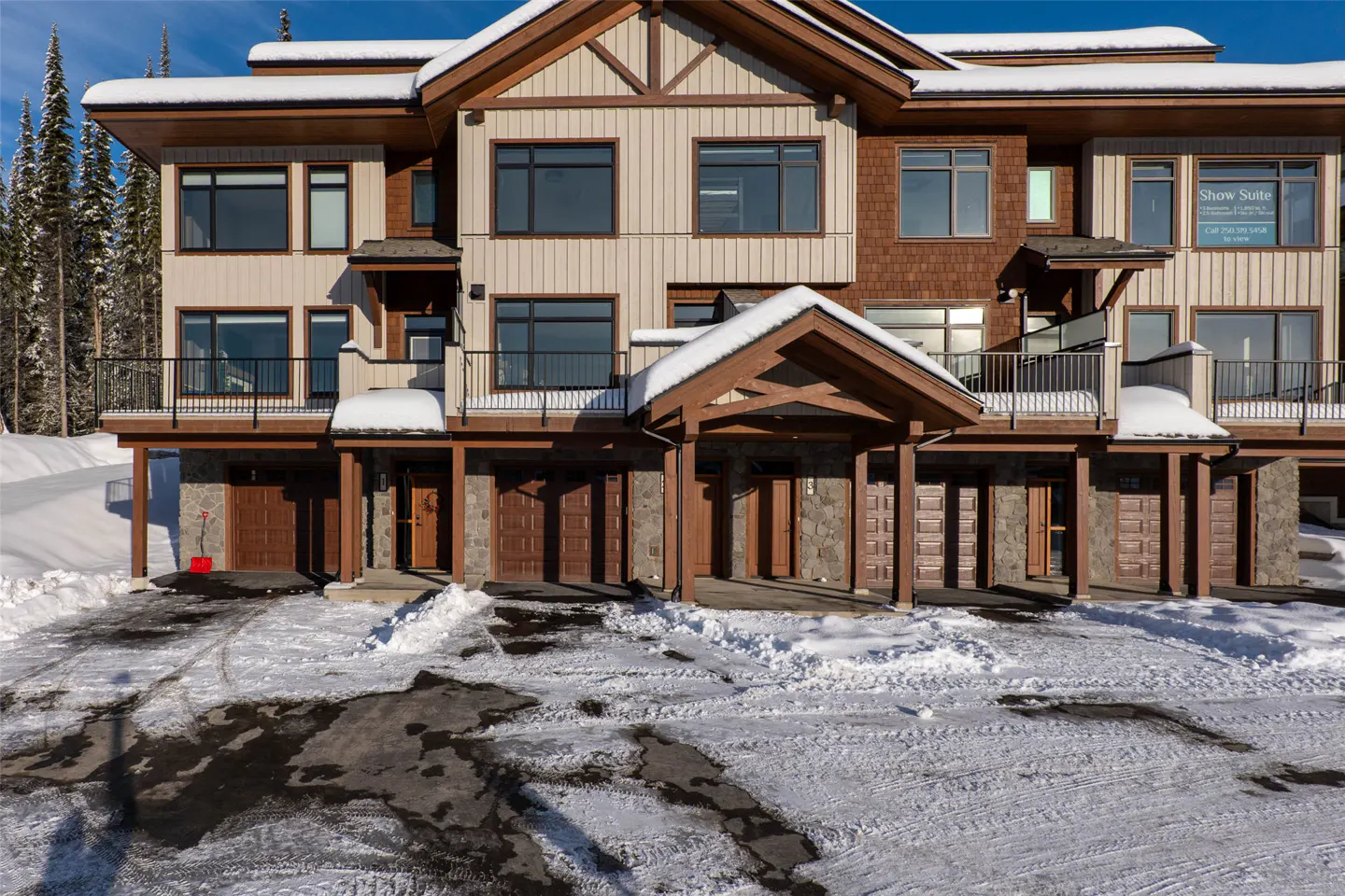 Three-story townhouse with brown trim, beige siding, and snow-covered roof. Each unit has a balcony and a garage. Snowy driveway in front.