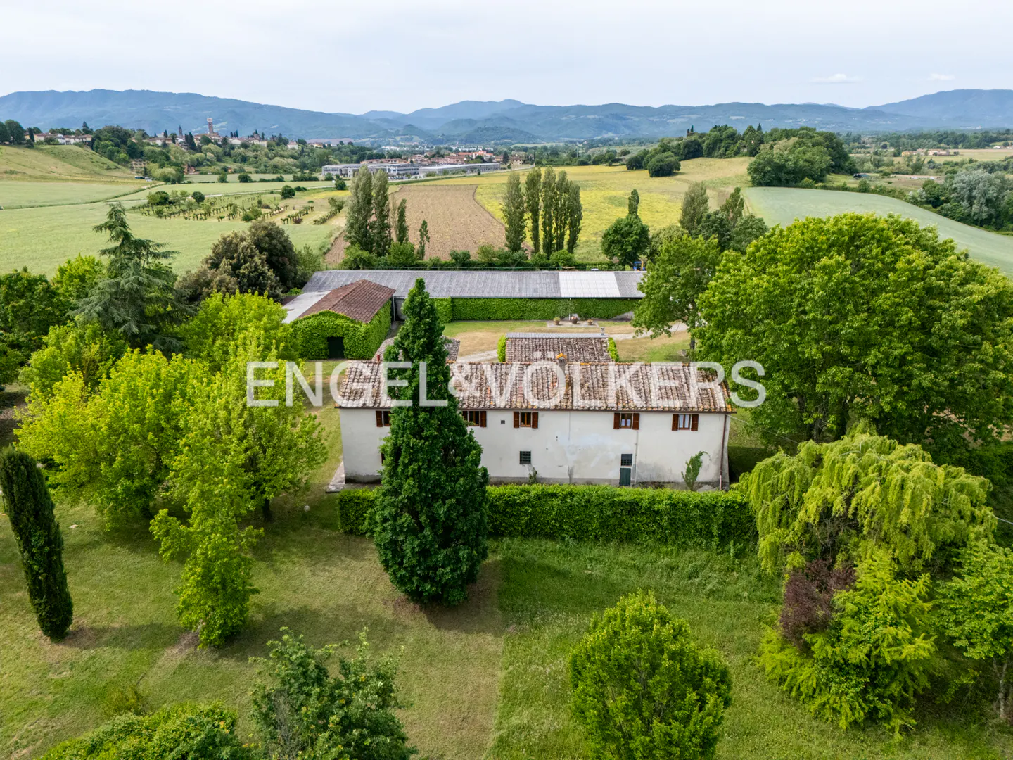 Aerial view of a white farmhouse with brown shutters, surrounded by green trees and fields, under a cloudy sky.