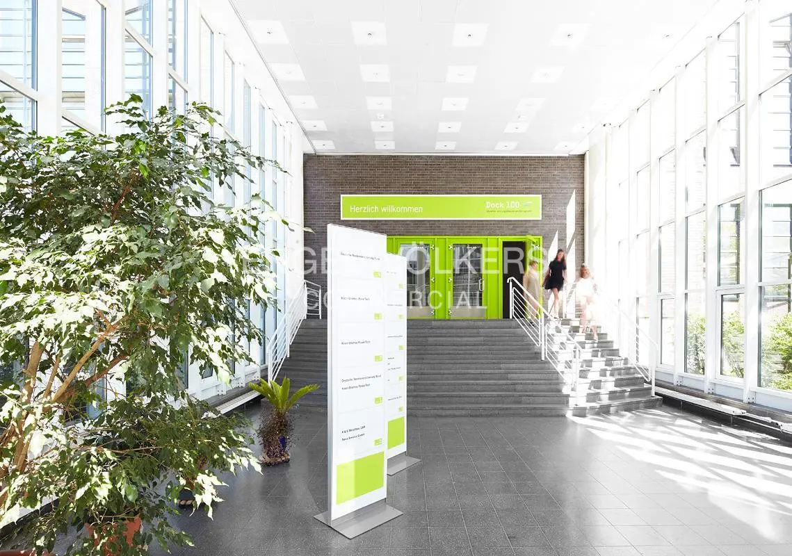 Bright lobby with large windows, stairs, and green doors. Two women walk down the stairs. A large plant is on the left.
