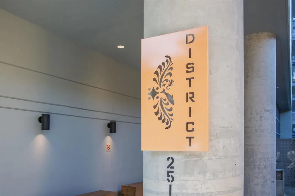 Exterior view of a building with a peach-colored sign reading "DISTRICT" with a floral design, mounted on a gray concrete pillar.