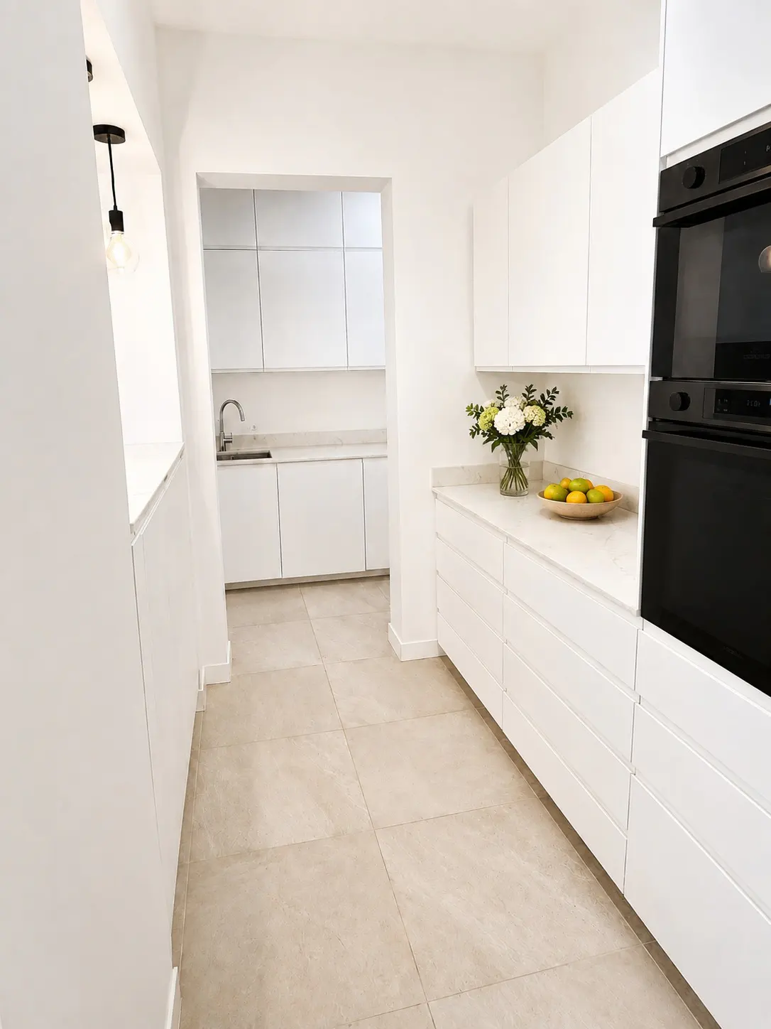 Bright, modern kitchen with white cabinets, countertops, and walls. A black oven is on the right, and a vase of flowers sits on the counter.