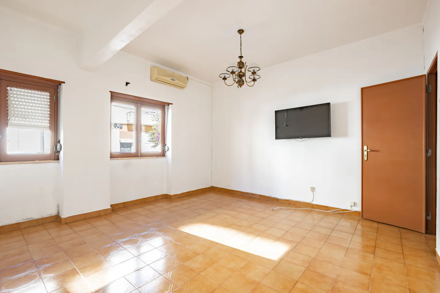 Bright, empty room with tile floor, white walls, and two windows. A TV hangs on the wall, and a chandelier hangs from the ceiling.