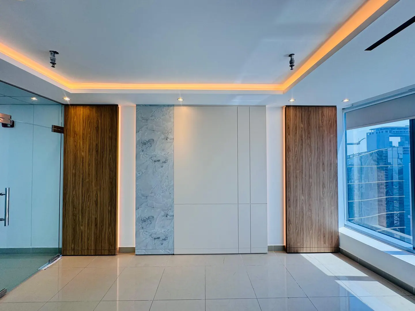 Empty office space with tile floors, a glass door, and a large window with a city view. The walls are white with wood and marble accents.